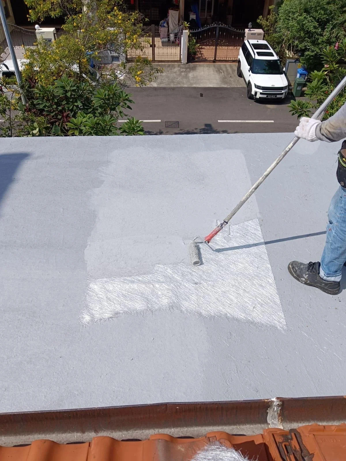 A person painting a flat roof with white waterproof coating, using a roller brush, on a sunny day.