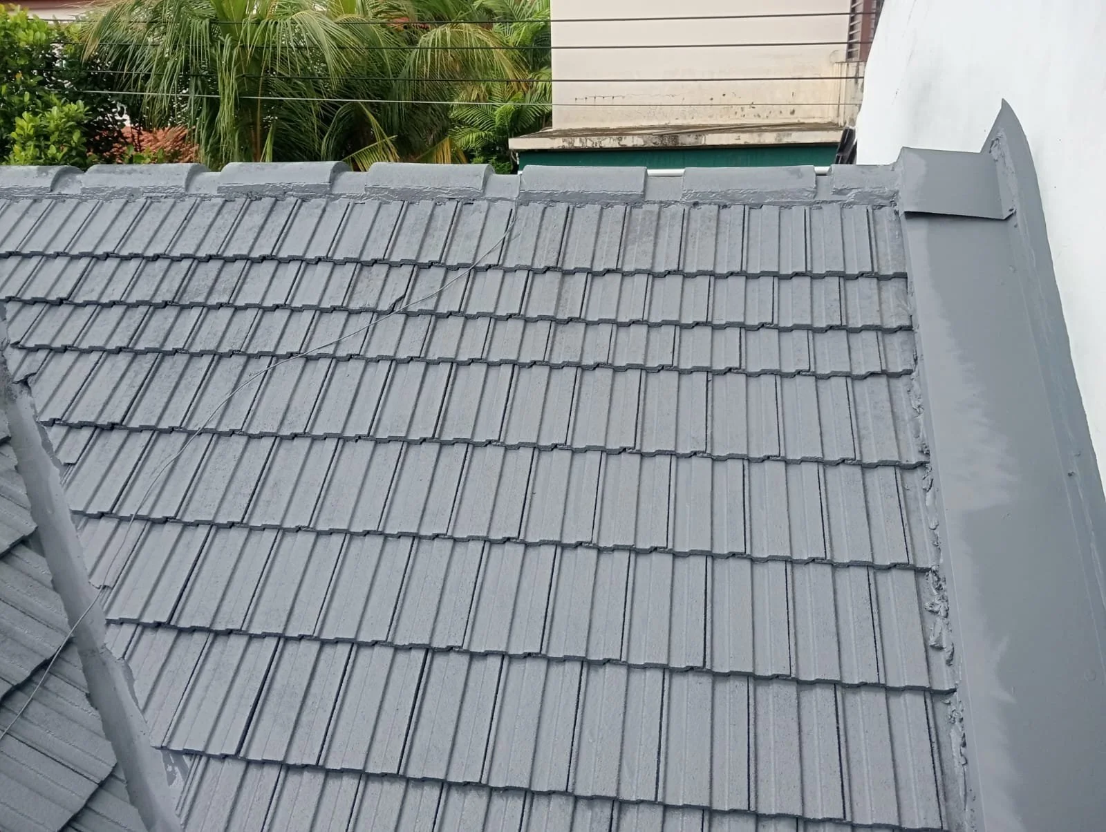 Close-up view of a roof with grey shingles and fresh grey sealant along the edges, with some trees and part of a building in the background.