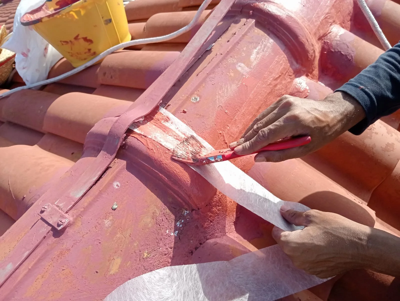 A person is painting or restoring decorative elements on a terracotta roof tile with a brush, using white paint or primer, in an outdoor setting.