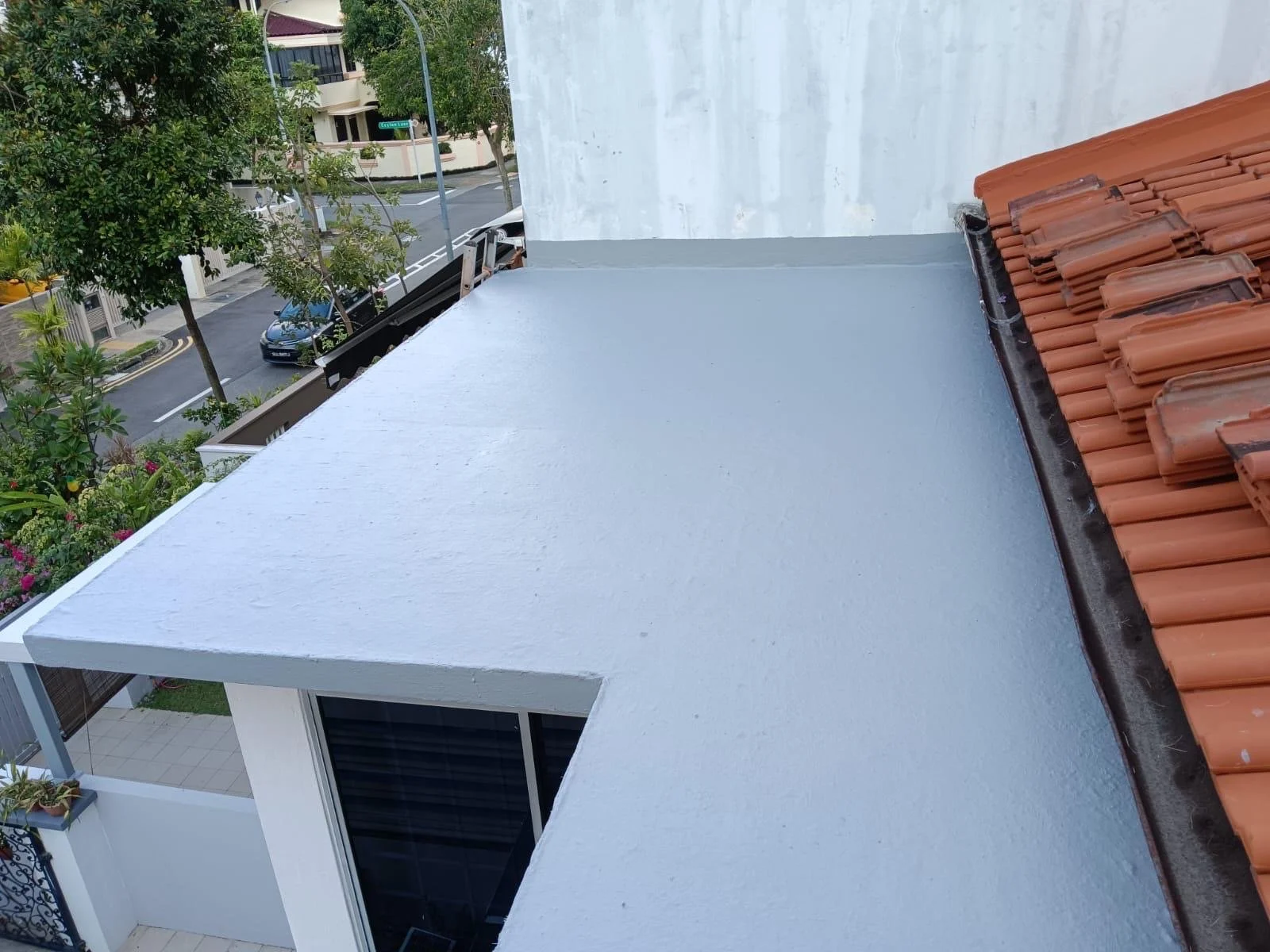View from a balcony showing a white rooftop extension of a building, red clay roof tiles, and a street with parked cars and trees in the background.