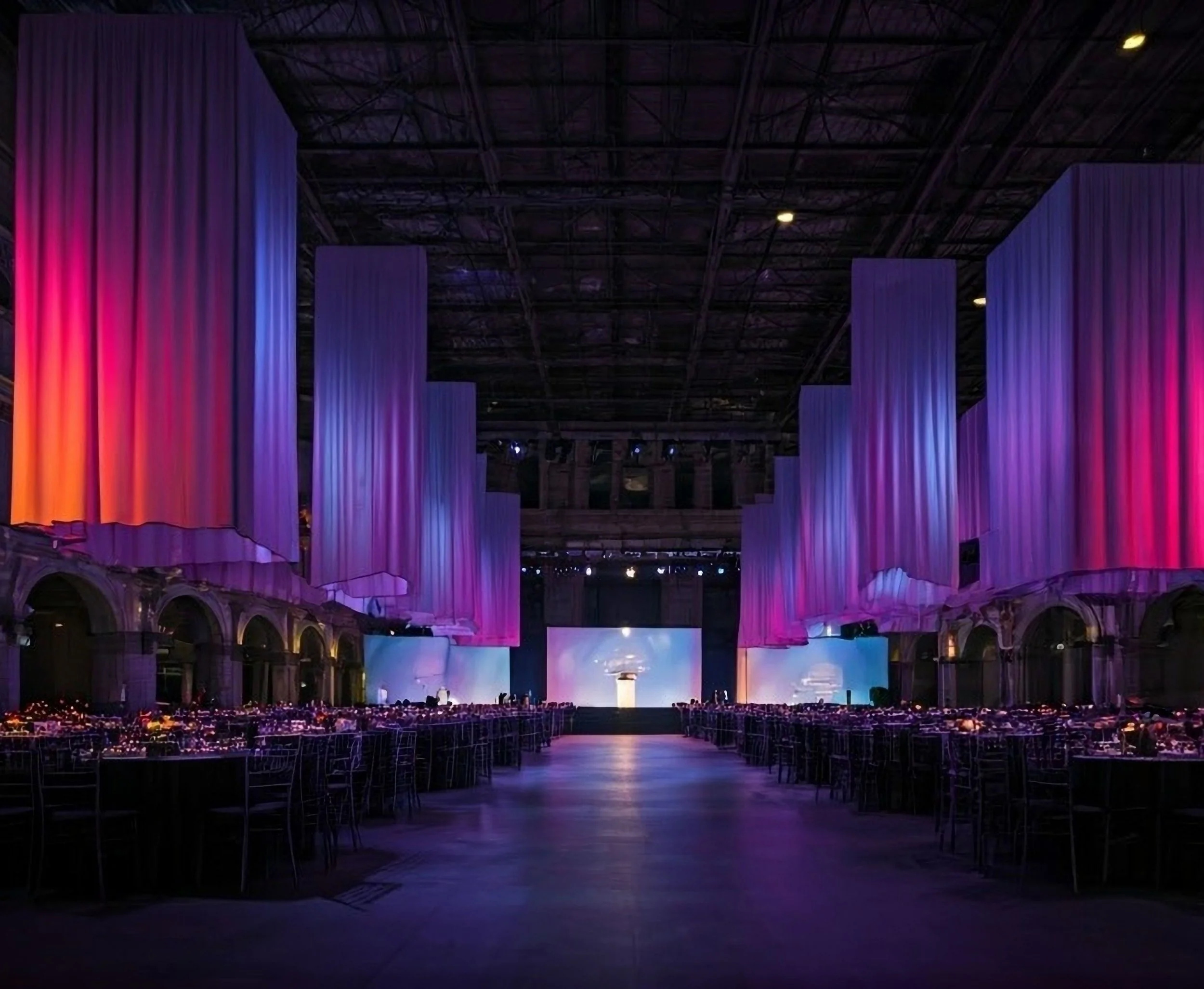 A large indoor event space decorated with multicolored lighting and draped curtains, with banquet tables and chairs set up for a formal gathering or celebration.