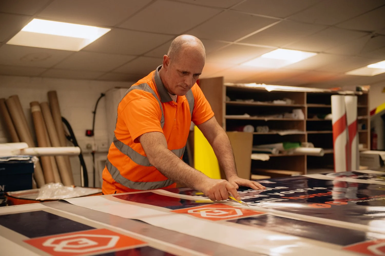 Worker in an orange safety shirt applying a sticker or decal to a large sign in a workshop.