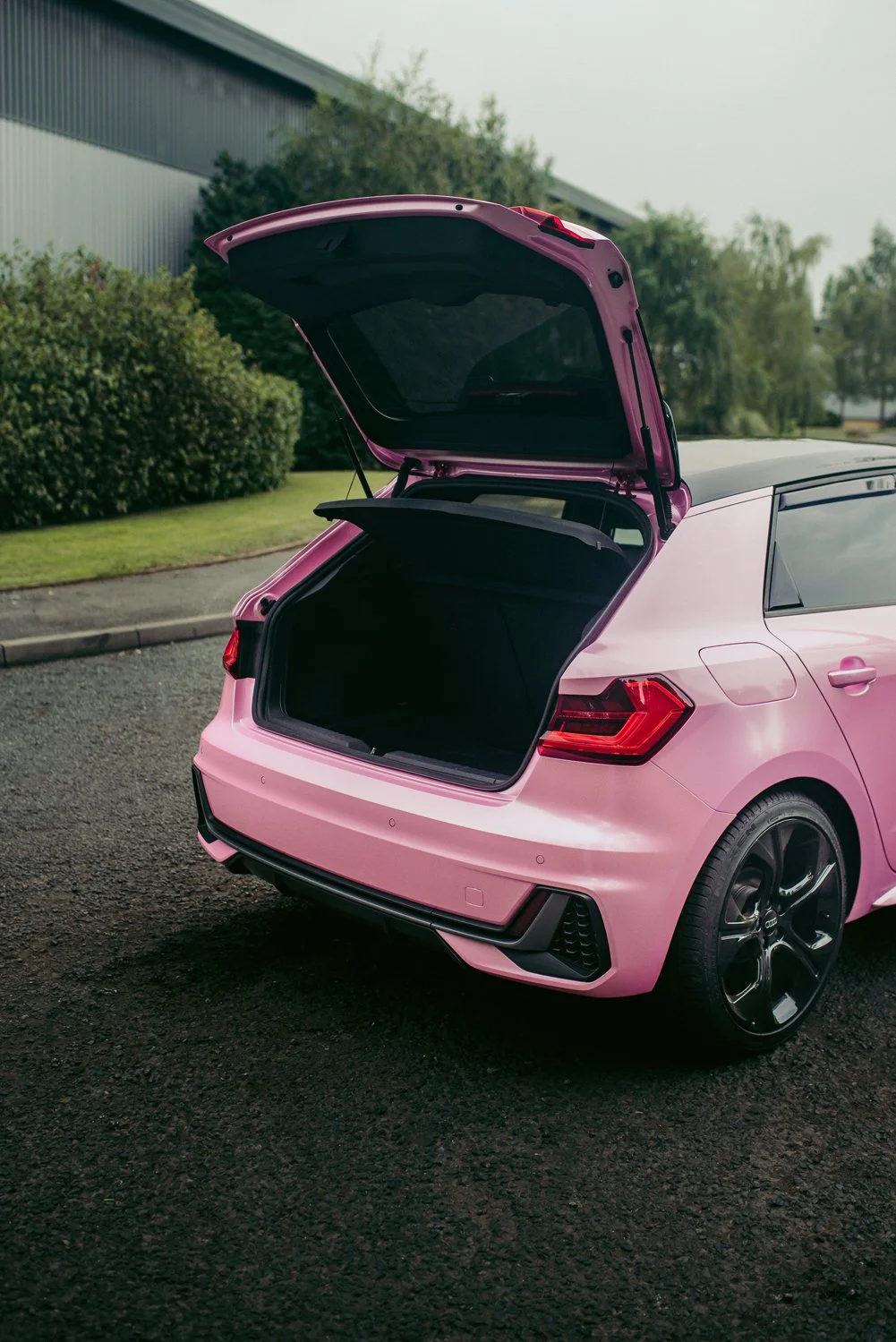 Pink hatchback car with open trunk parked on a paved surface near green bushes and trees.