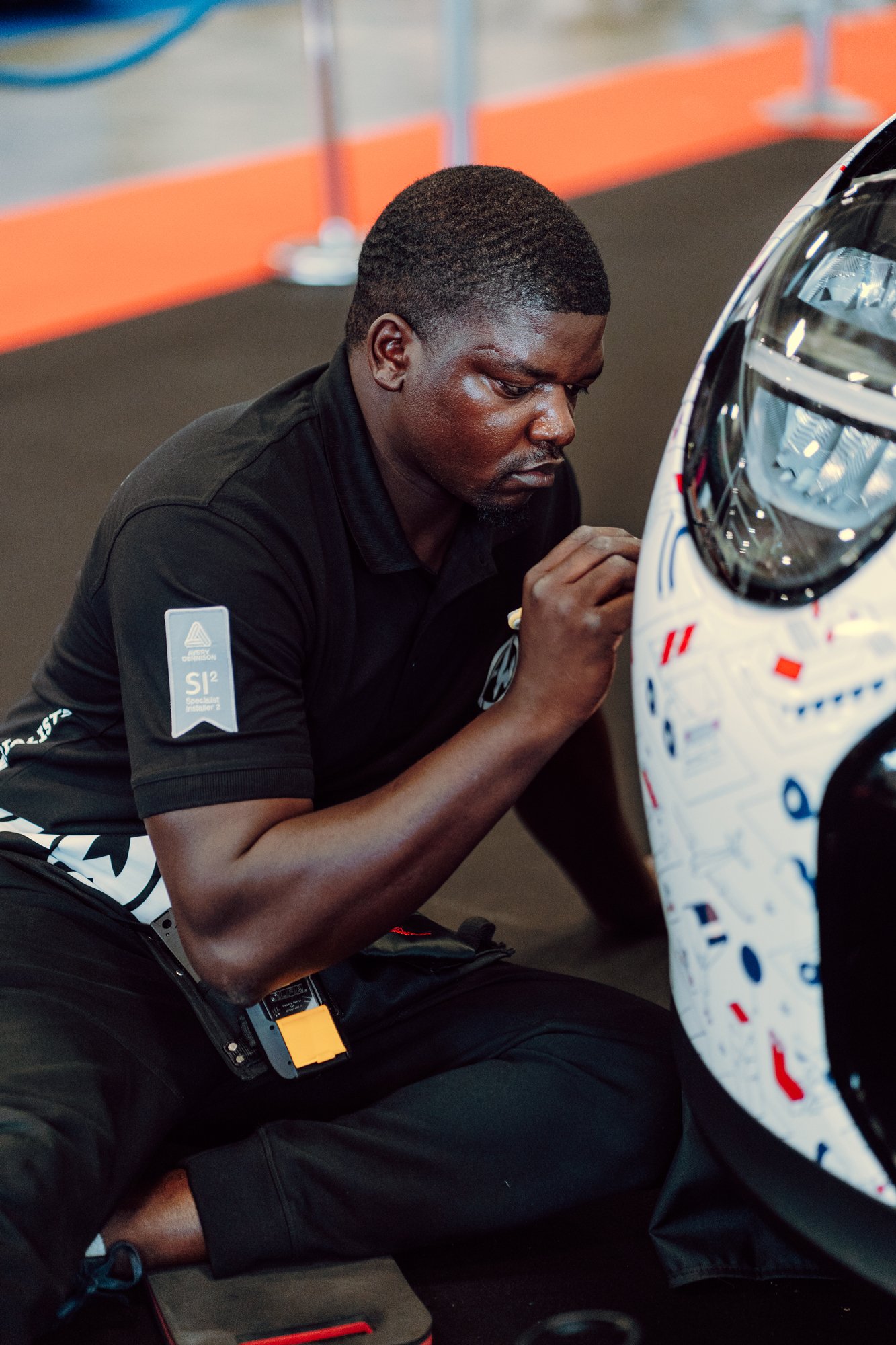 A man in a black shirt working on a race car, crouched beside it in a garage or workshop environment.