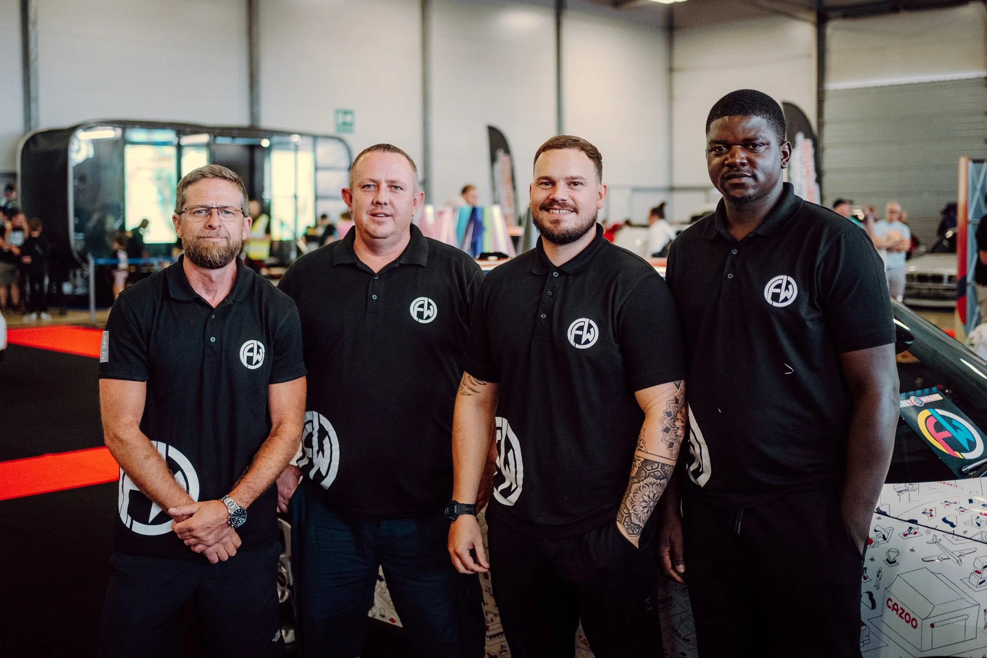 Four men wearing black polo shirts with a white logo, standing indoors at an event, with people and booths visible in the background.