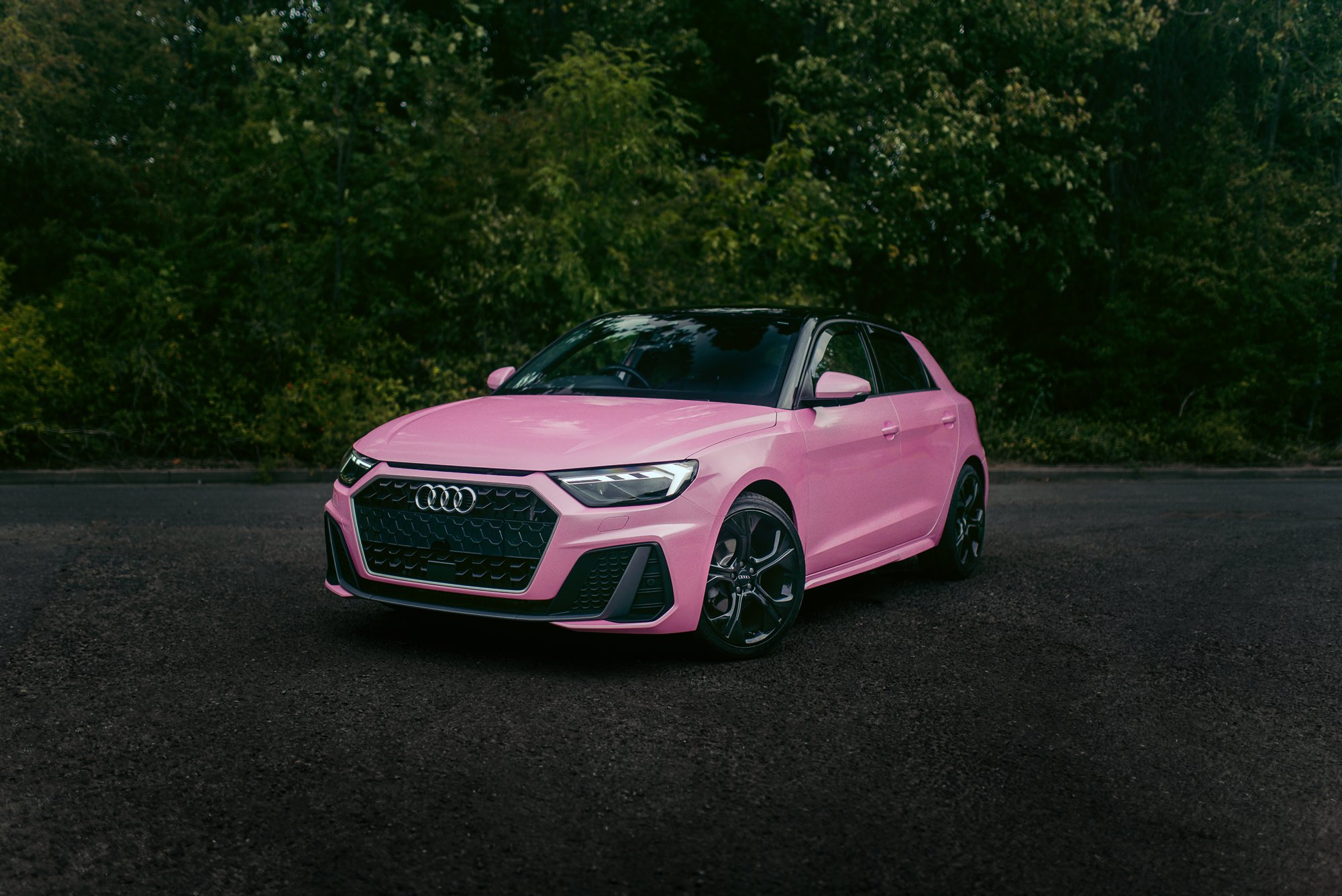 Pink Audi hatchback car parked on a dark paved road with green trees in the background.