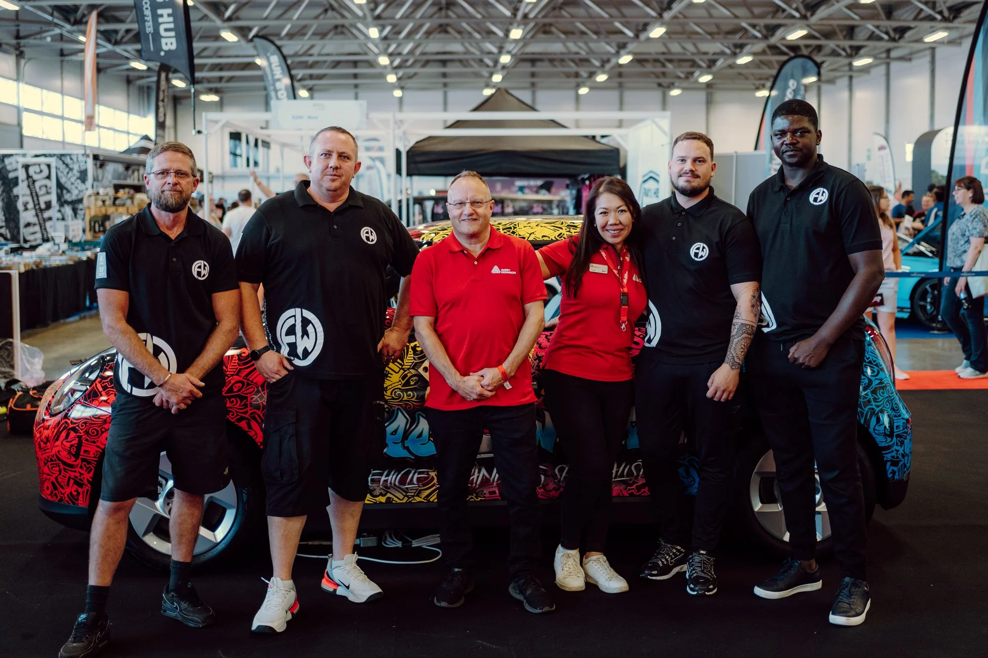 A group of six people standing in front of a colorful car at a trade show or exhibition. The background shows booths and other attendees.