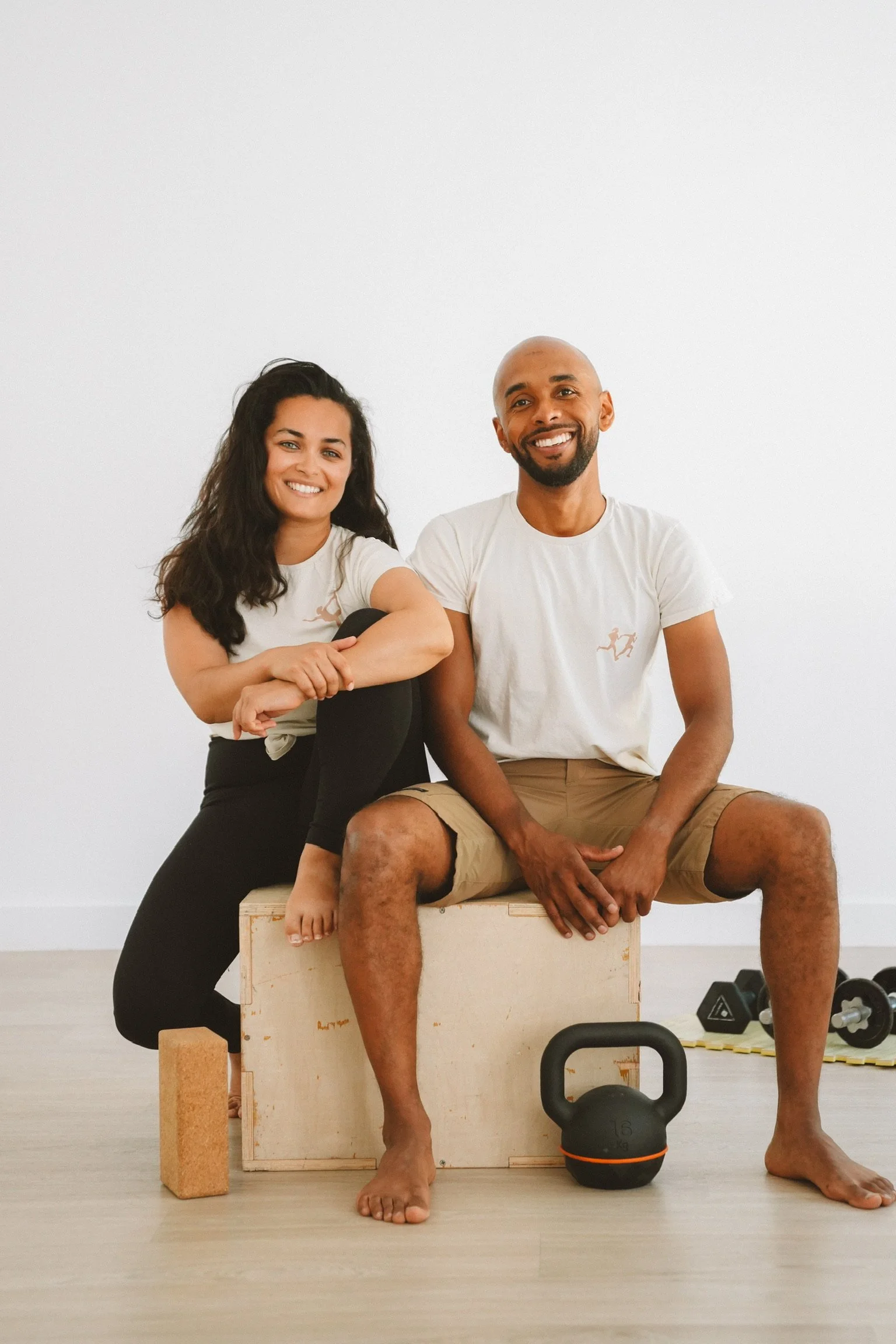 Deux personnes assises, domestiquées et souriantes dans une salle de sport, avec des haltères et un kettlebell au sol, un tapis de yoga, un casse-cou et un mur blanc en arrière-plan.