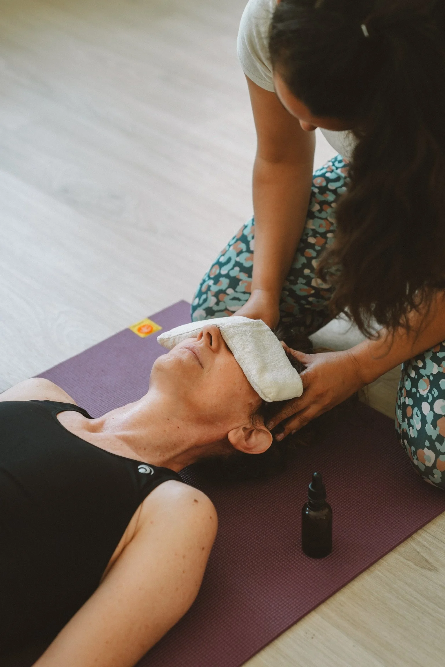 Une femme effectue une séance de thérapie ou de massage sur une autre personne allongée sur un tapis de yoga, en utilisant de l'huile essentielle dans une bouteille opaque.