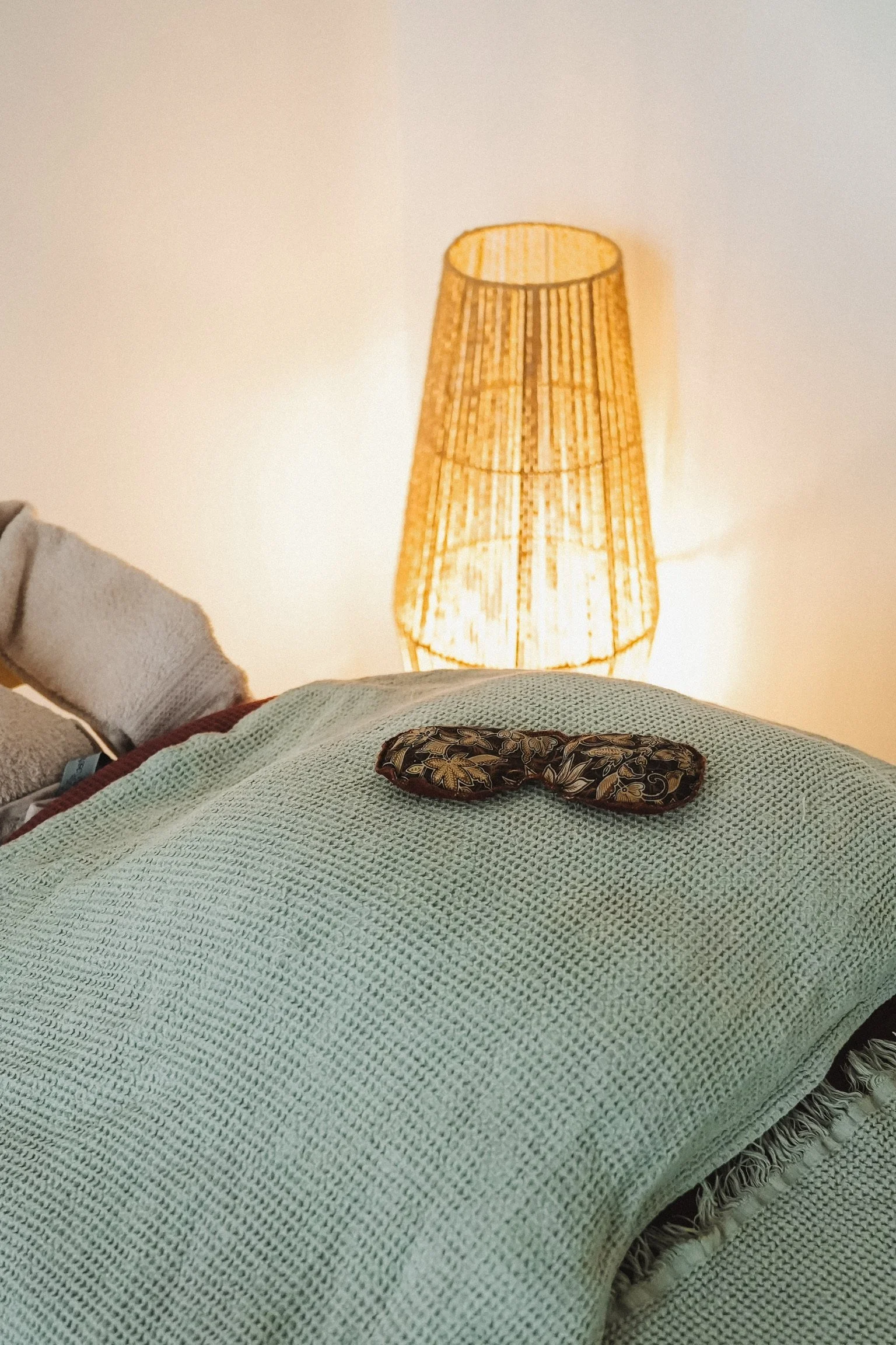 L'image montre une lampe en osier sur une table, une couverture en tricot vert et un masque de sommeil avec un motif floral. Un côté du lit est visible, avec un coussin coloré au coin.