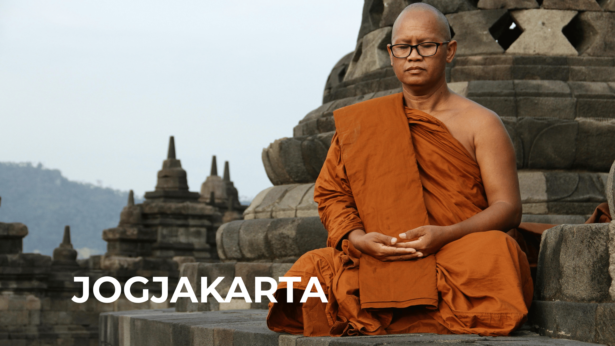 A Buddhist monk in orange robes sitting in meditation near ancient stone temples in Yogyakarta, Indonesia.
