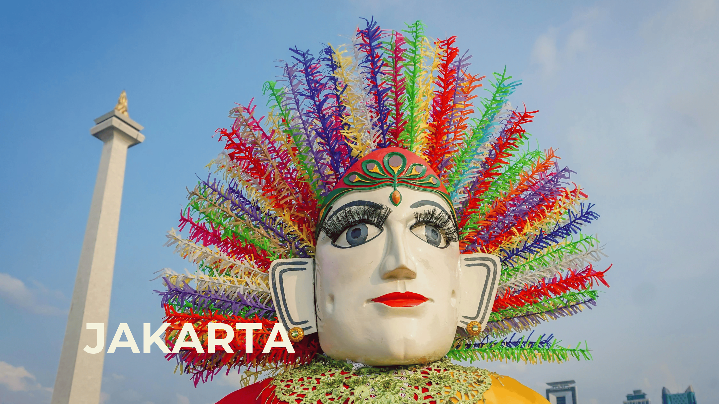 A large decorative mask of a woman with an elaborate multicolored headdress made of plastic or paper, set against a blue sky with a tall monument in the background.