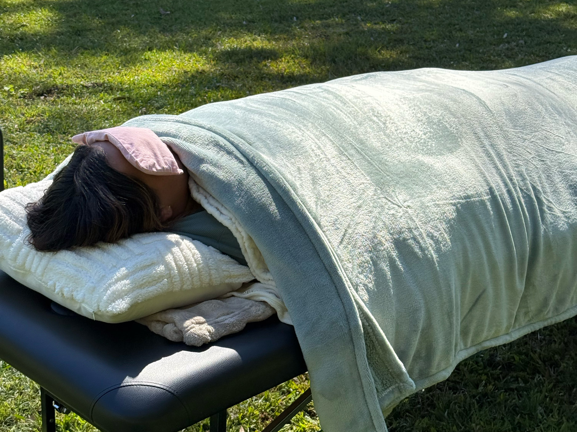 Person lying on a massage table outdoors, covered with a light-colored blanket, resting with a pink eye mask, under sunlight.