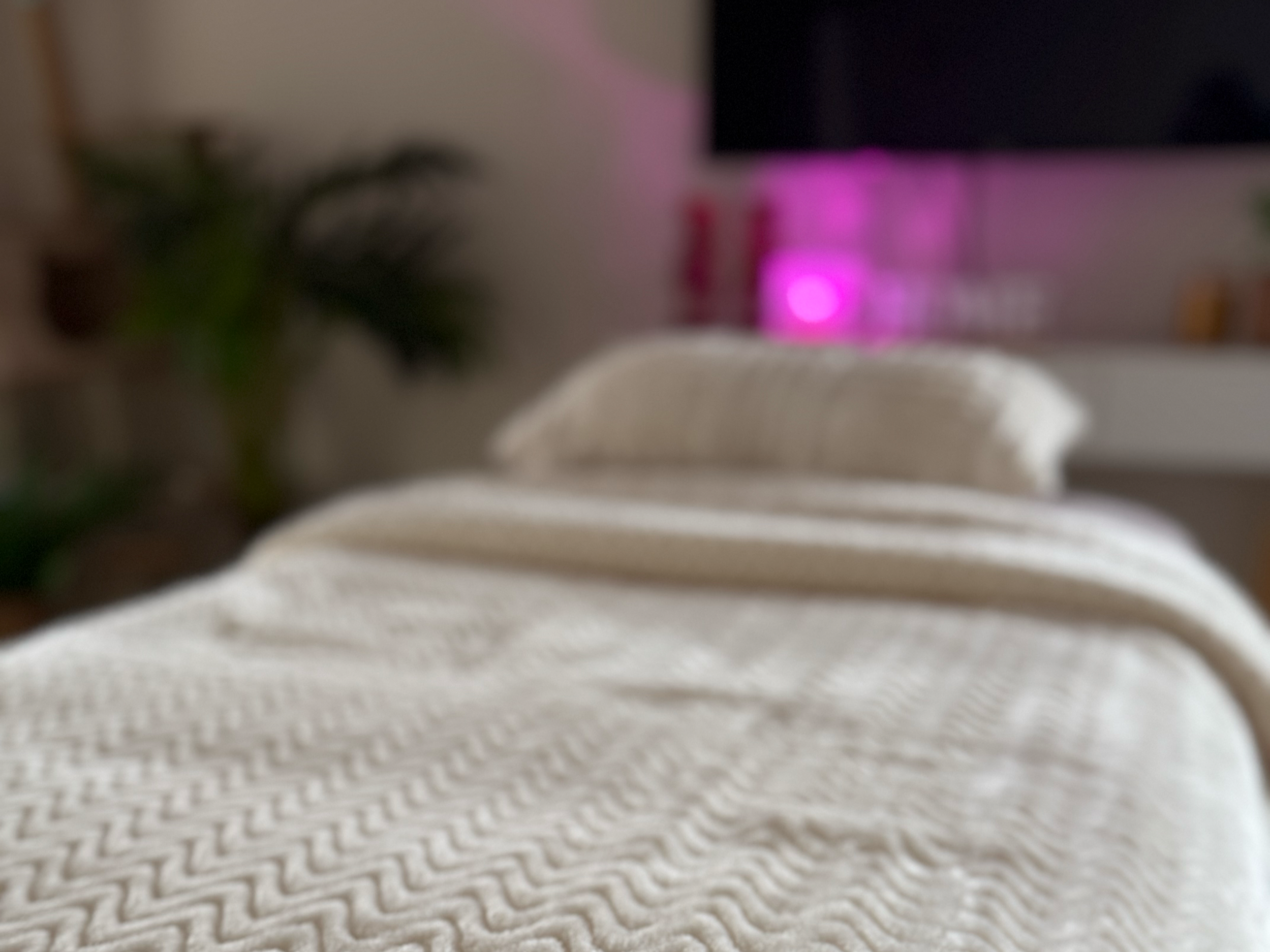 Close-up of a fluffy white blanket on a bed with pillows, a TV, and a pink-lit room in the background.