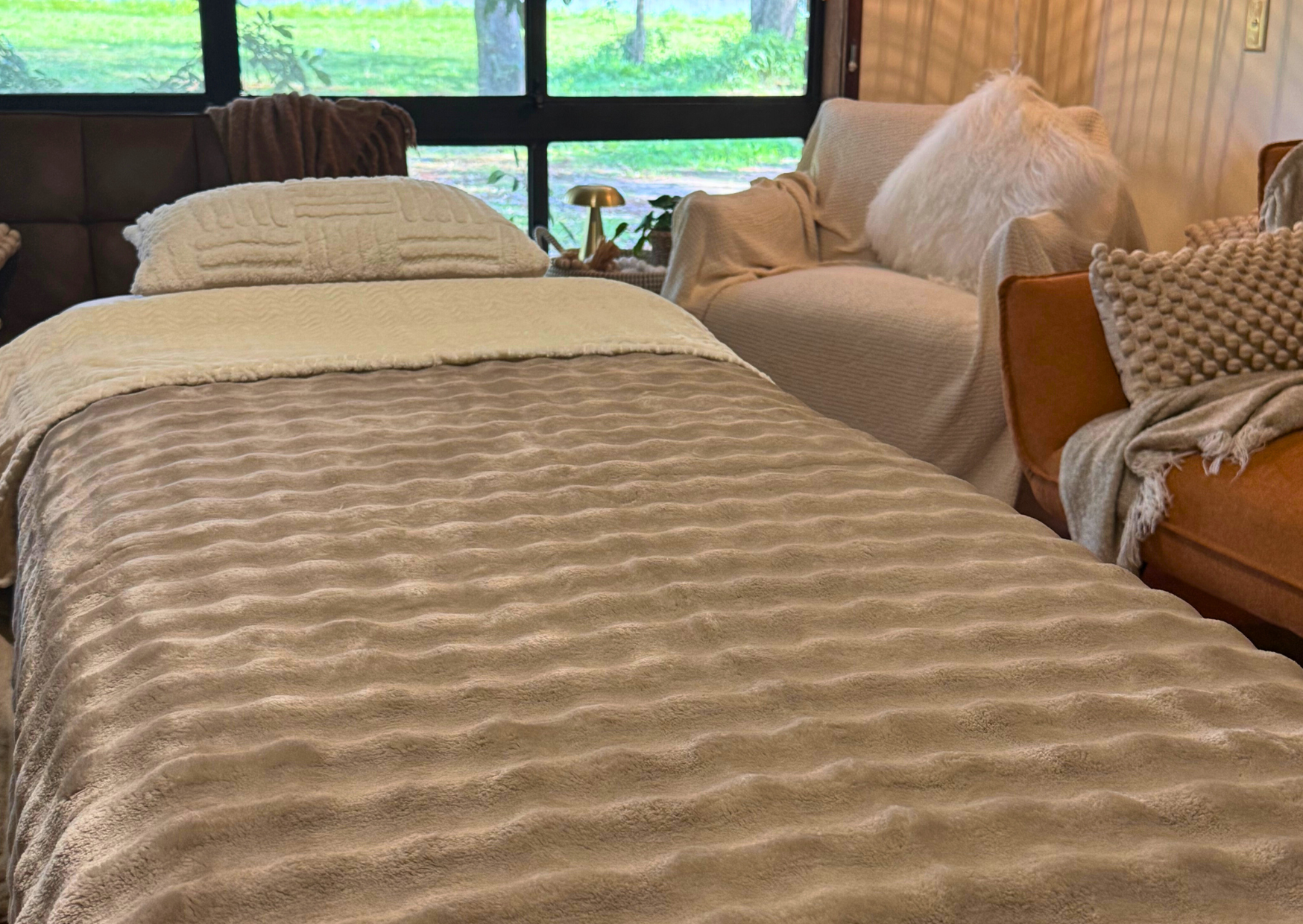 A bed with beige textured bedding and pillows in a cozy room with large windows and outdoor greenery in the background, flanked by two sofas with blankets and pillows.