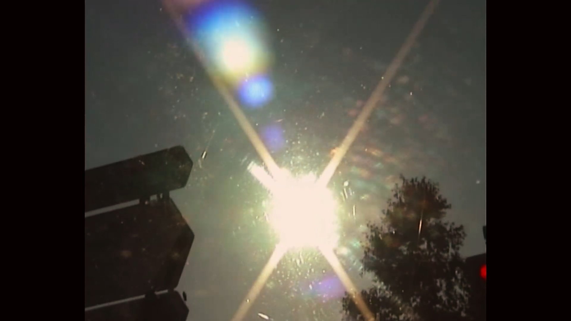 Bright fireworks explosion seen through a windshield at night with tree silhouettes and vehicle parts visible.