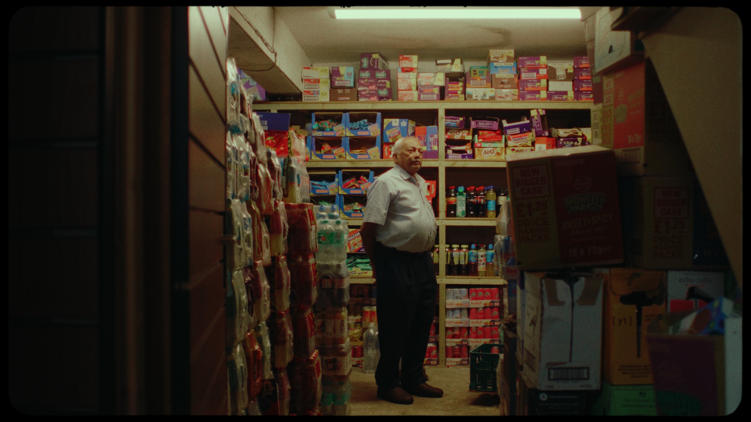 An older man stands inside a small grocery or convenience store filled with shelves of snacks, drinks, and food products. The man is looking to the side, wearing a light-colored shirt and dark pants.