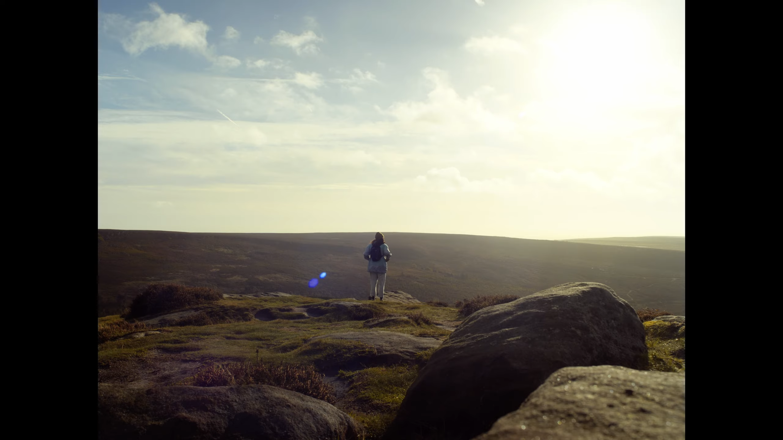 A person standing on a grassy hilltop, looking towards a vast open landscape with rolling hills under a partly cloudy sky, sunlight shining from the right side.
