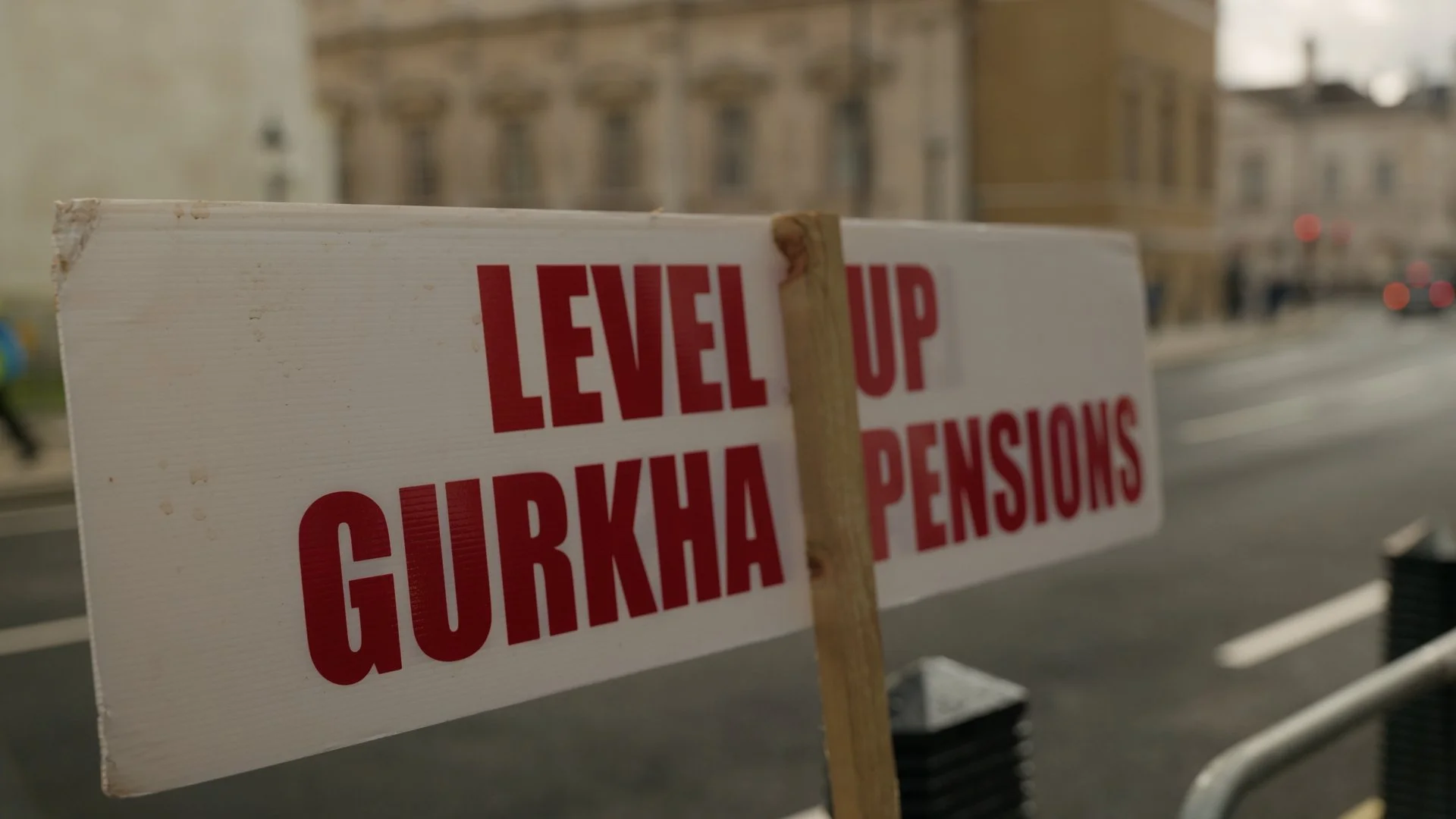 A white sign with red text that reads 'LEVEL UP GURKHA PENSIONS' on a city street.