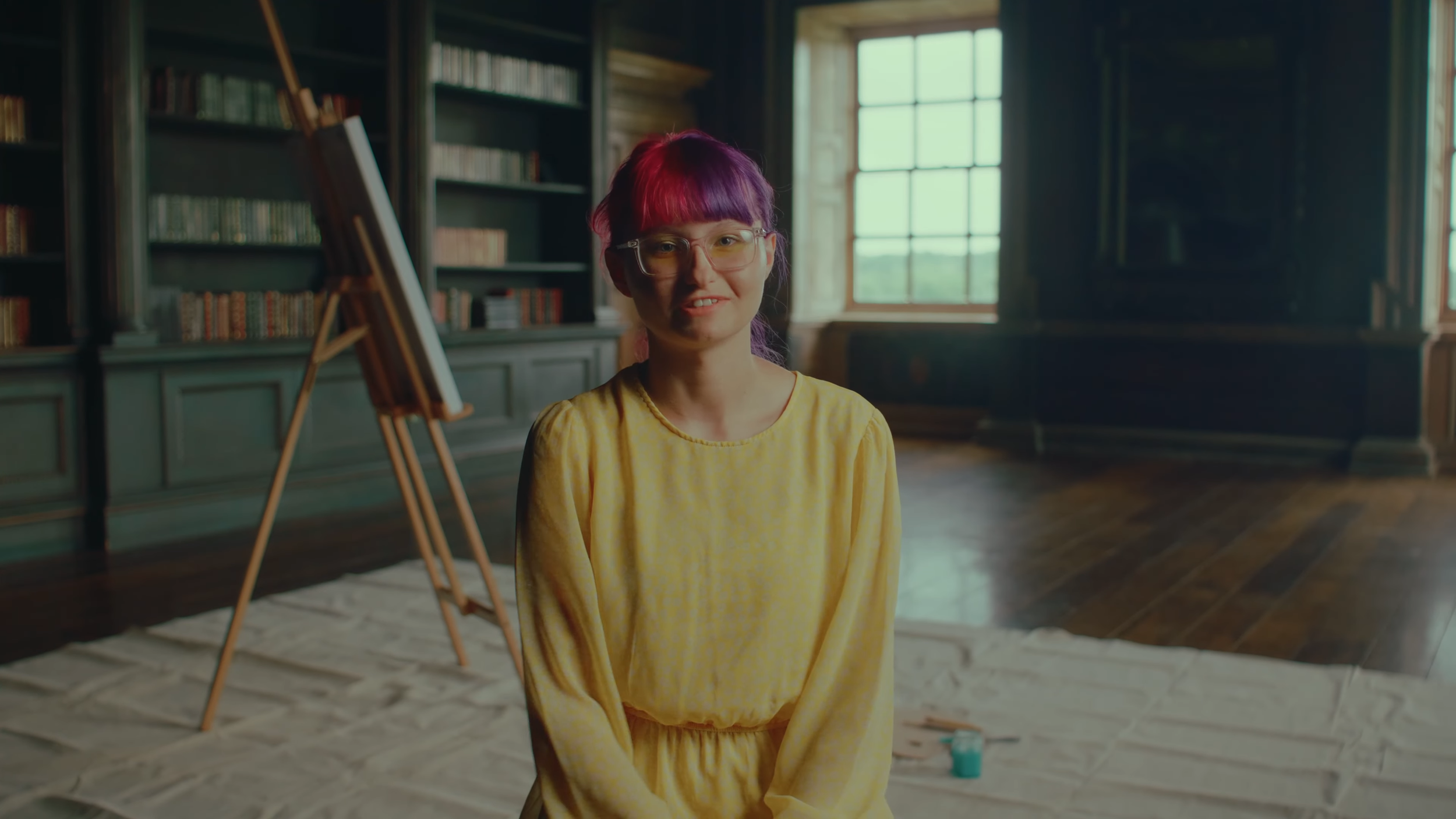 A woman with pink and purple hair and glasses sits in a room with dark green bookshelves, large windows, and wooden flooring, with an easel and art supplies nearby.