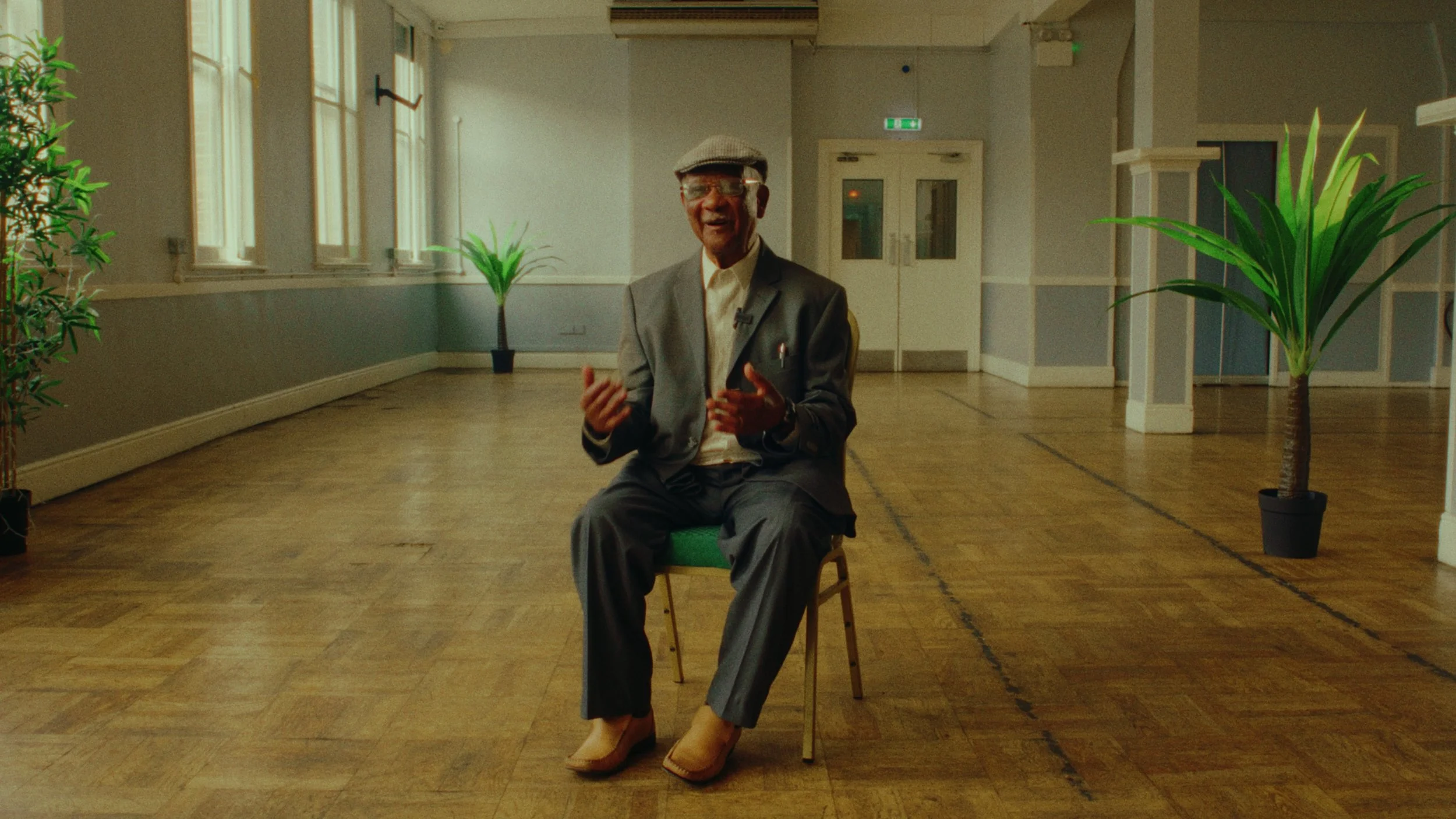 An elderly man in a gray suit and hat sitting on a green chair in a spacious, empty room with wooden floors and large windows, smiling and gesturing with his hands.