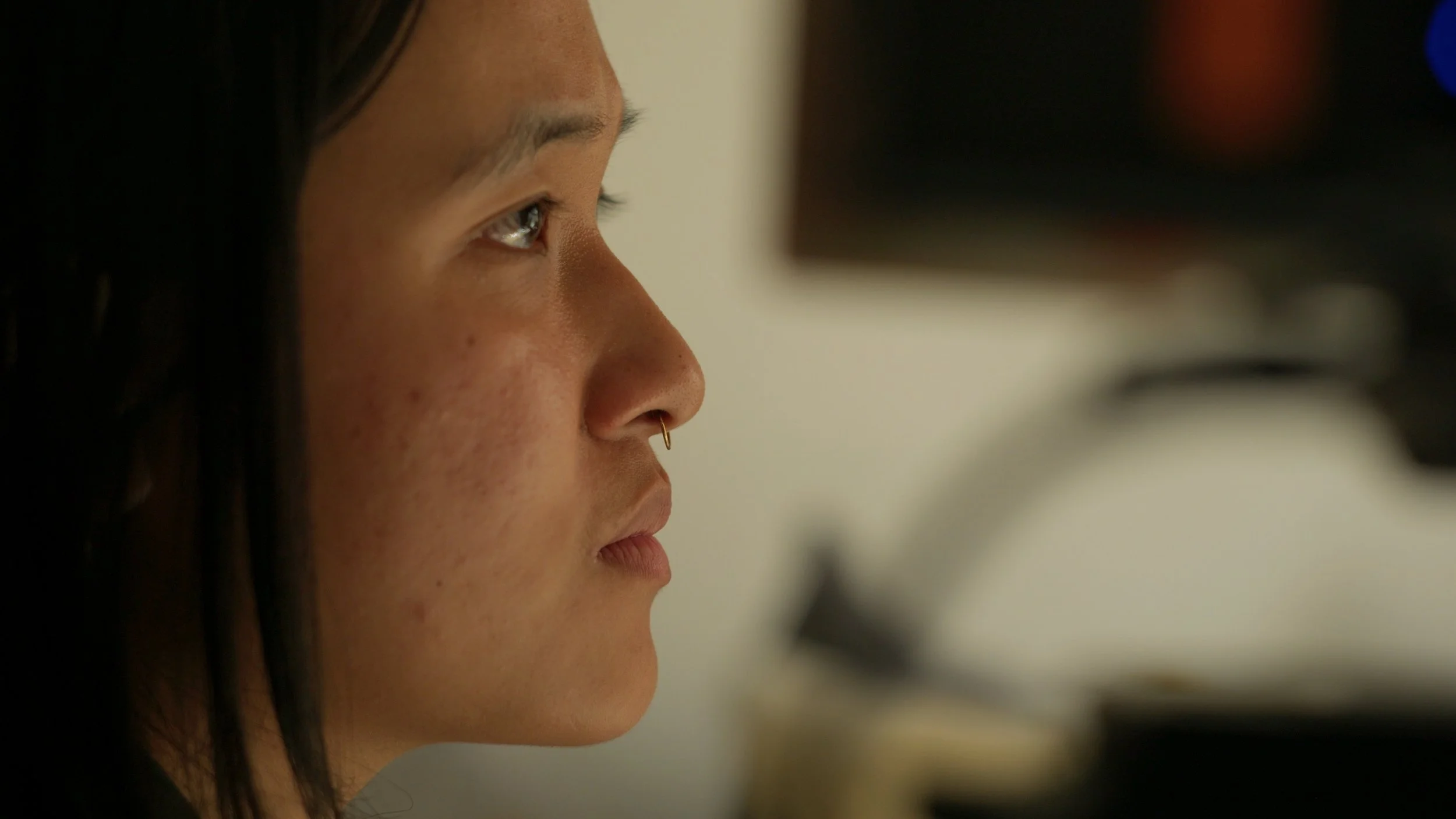 Close-up side profile of a person with dark hair, a septum piercing, and visible acne scars, looking intently at a computer screen.