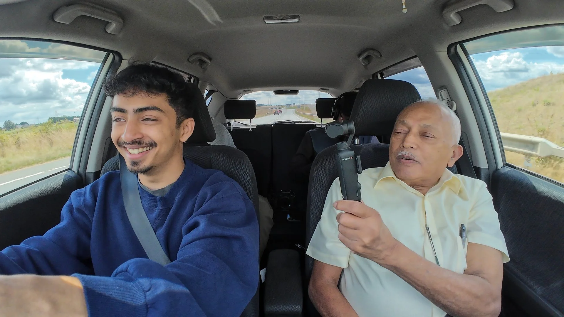 A young man driving in a car with an older man sitting next to him holding a device, both looking out the window on a highway with a grassy landscape and blue sky with clouds.