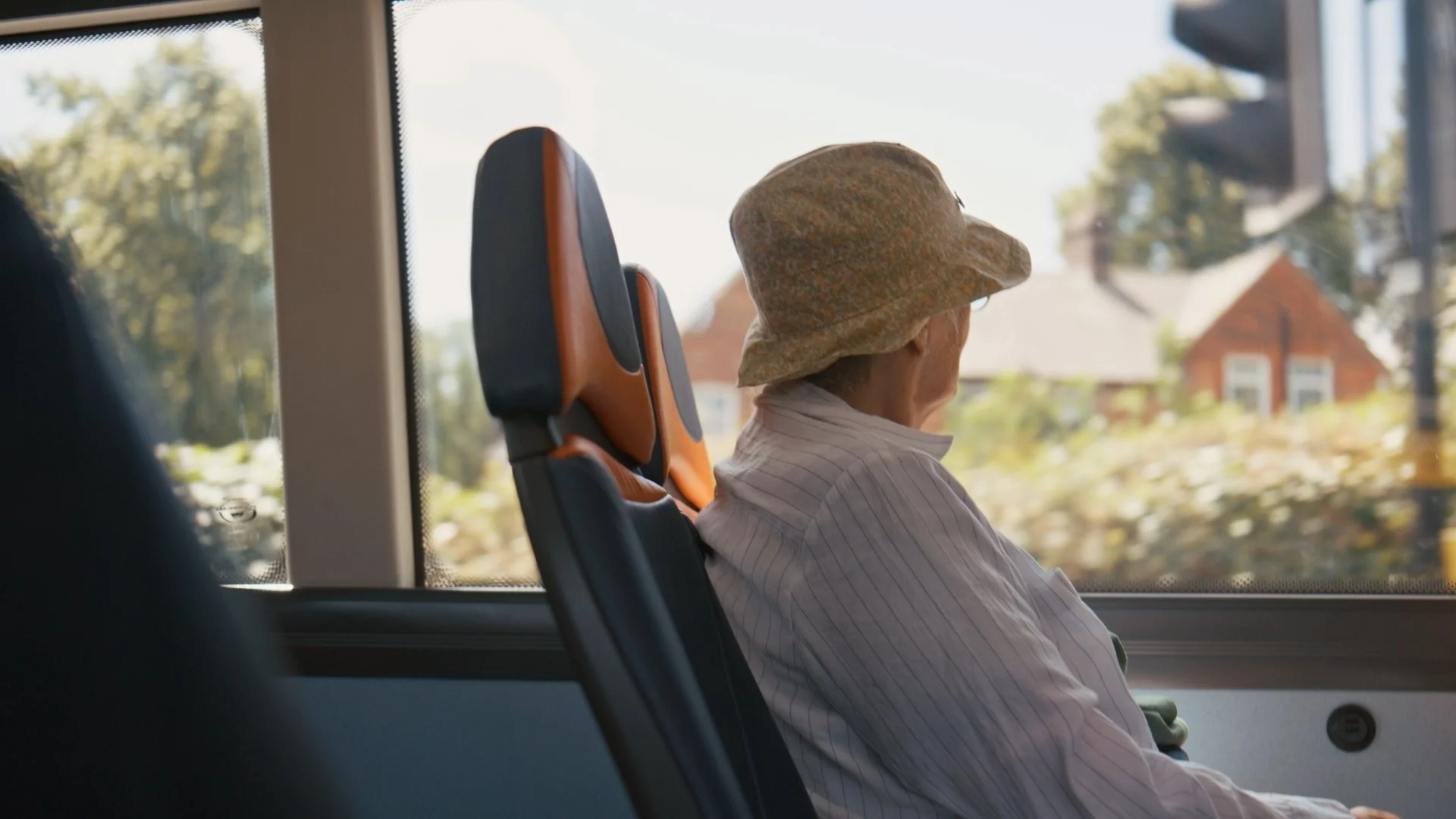 An elderly person wearing a hat and sitting on a bus is looking out the window at a rural landscape with houses and trees.