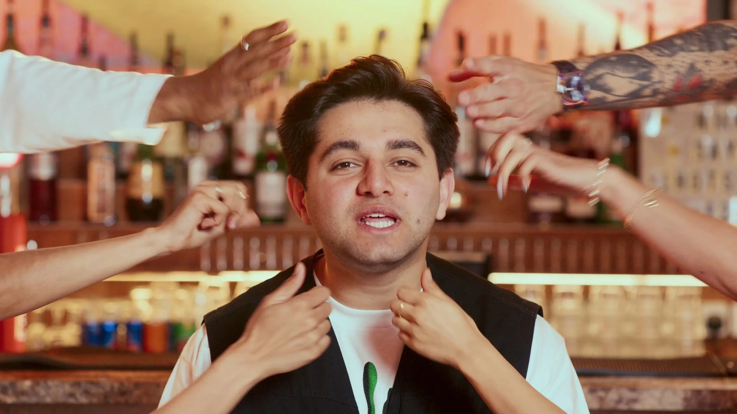A young man with dark hair sitting in a bar, surrounded by several hands reaching towards his head and neck, as if touching or adjusting his hair and shirt.