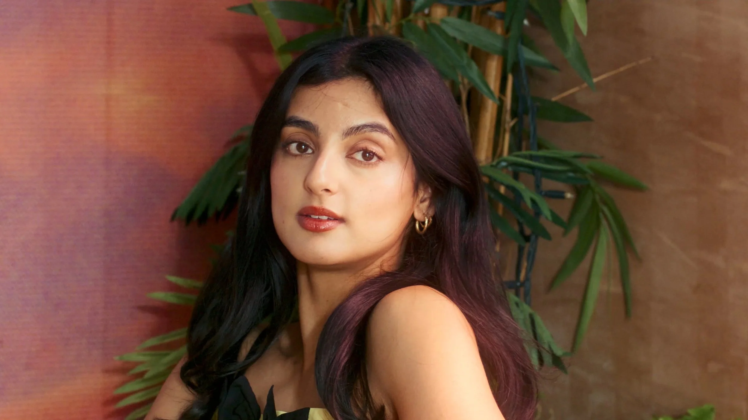 A woman with long dark hair and light makeup looking at the camera, with a wooden wall and green plant in the background.