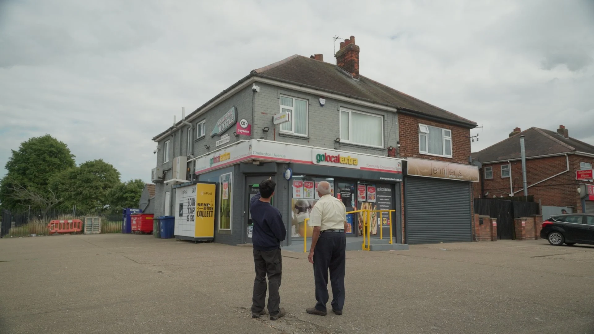 Two men standing outside a convenience store in a small parking lot on a cloudy day.