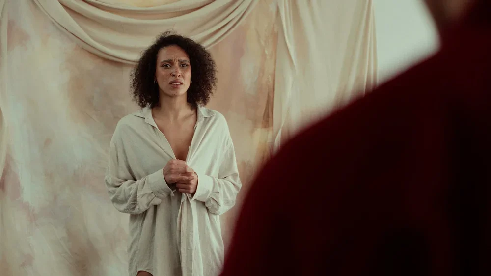 A woman with curly hair standing in front of a beige backdrop, looking distressed, with a person partially visible in the foreground.