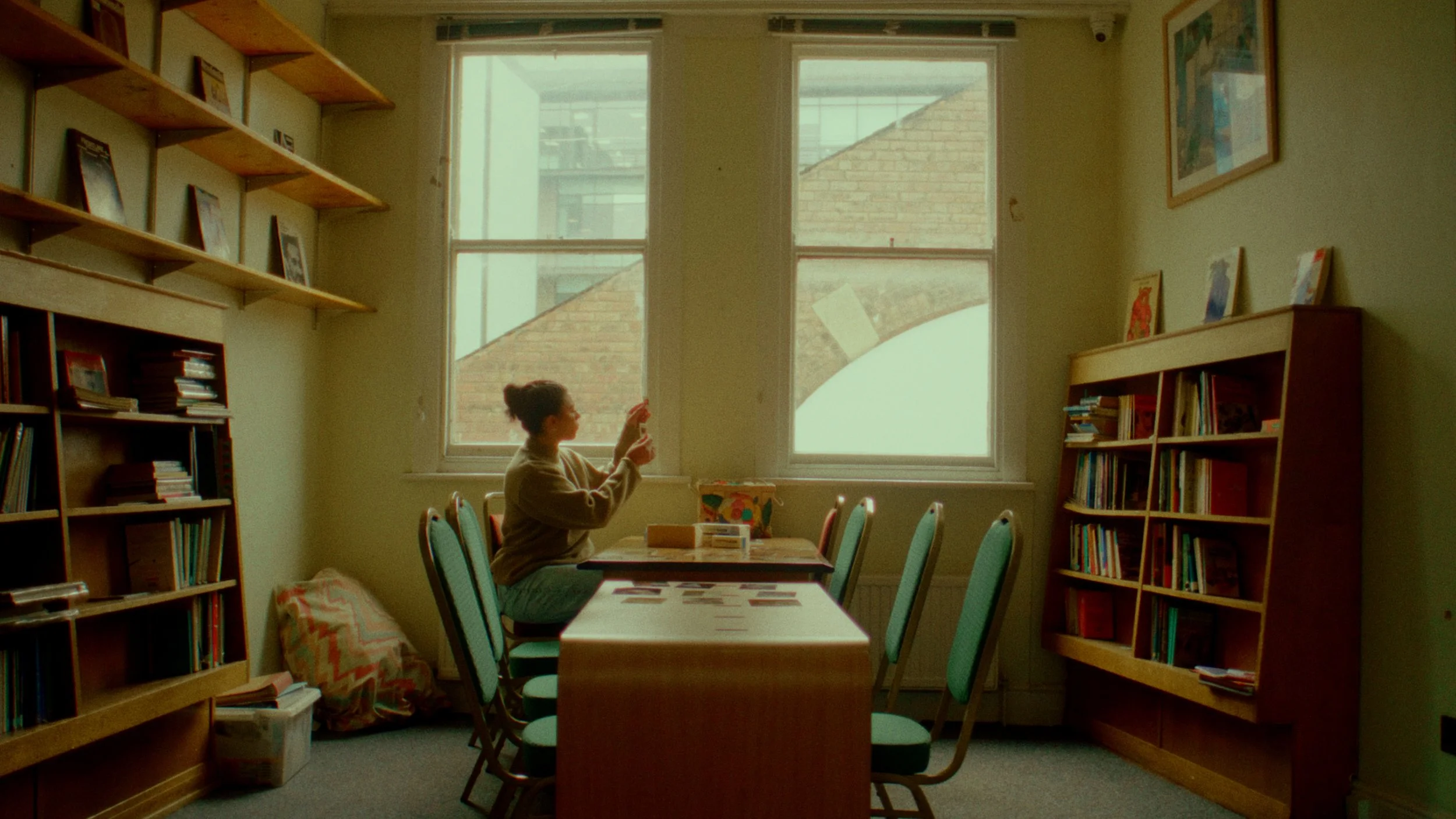 A woman sitting on a chair at a dining table inside a room with large windows, bookshelves, and on the table are various items.