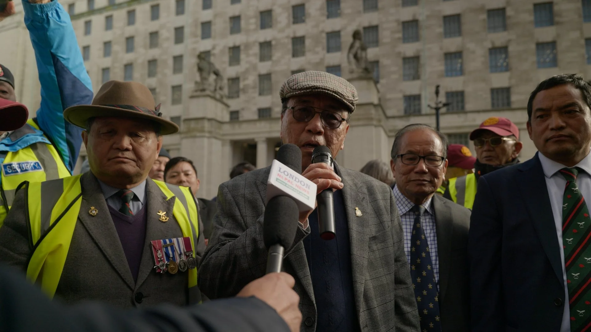 A group of men, including some with medals and badges, standing outdoors in front of a government building. One man is speaking into microphones held by reporters.