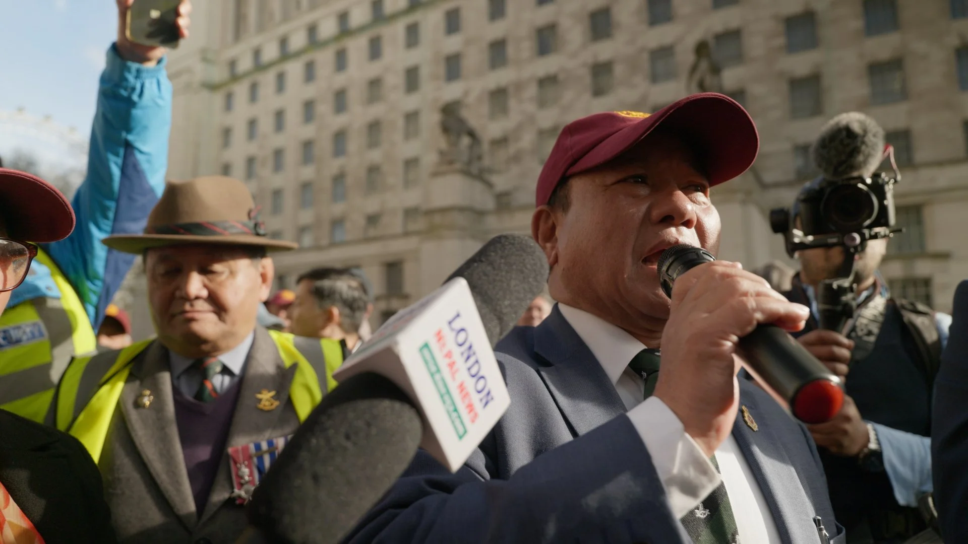 A man speaking into a microphone during a protest or rally, surrounded by people including a man in a military uniform and another in a high visibility vest, with a building in the background.