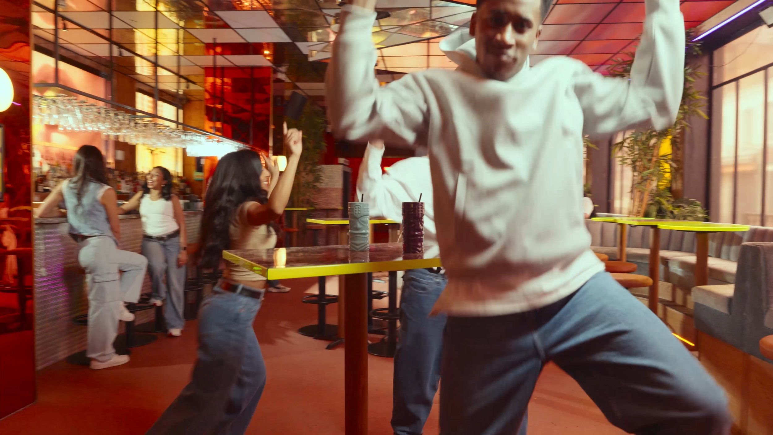 People dancing and socializing in a vibrant bar or lounge with colorful lighting and large windows.