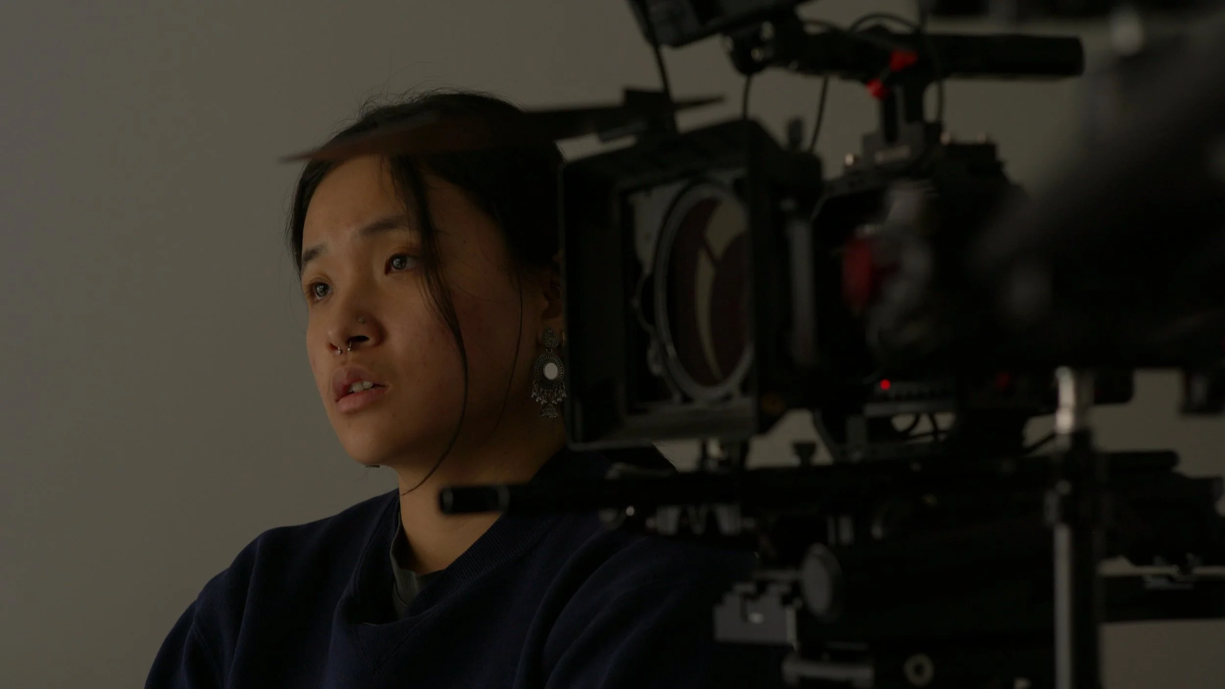 A woman with dark hair, nose piercing, and earrings sits in front of a film camera, looking thoughtfully to the side.