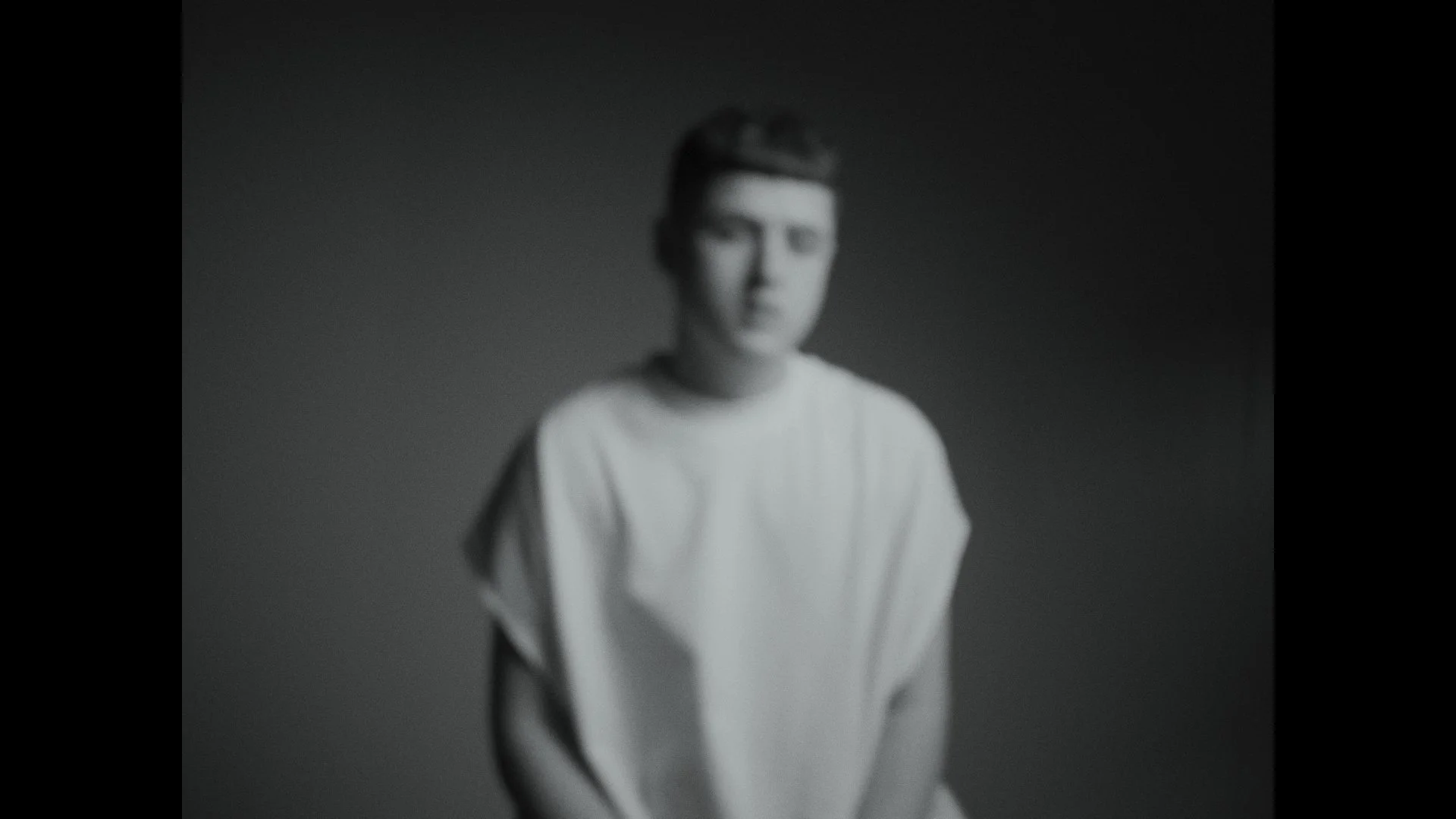 A blurred black and white photo of a person with short hair, wearing a light-colored top, standing in front of a dark background.