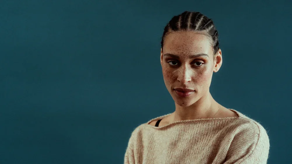 A woman with freckles in a beige sweater looking at the camera against a teal background.