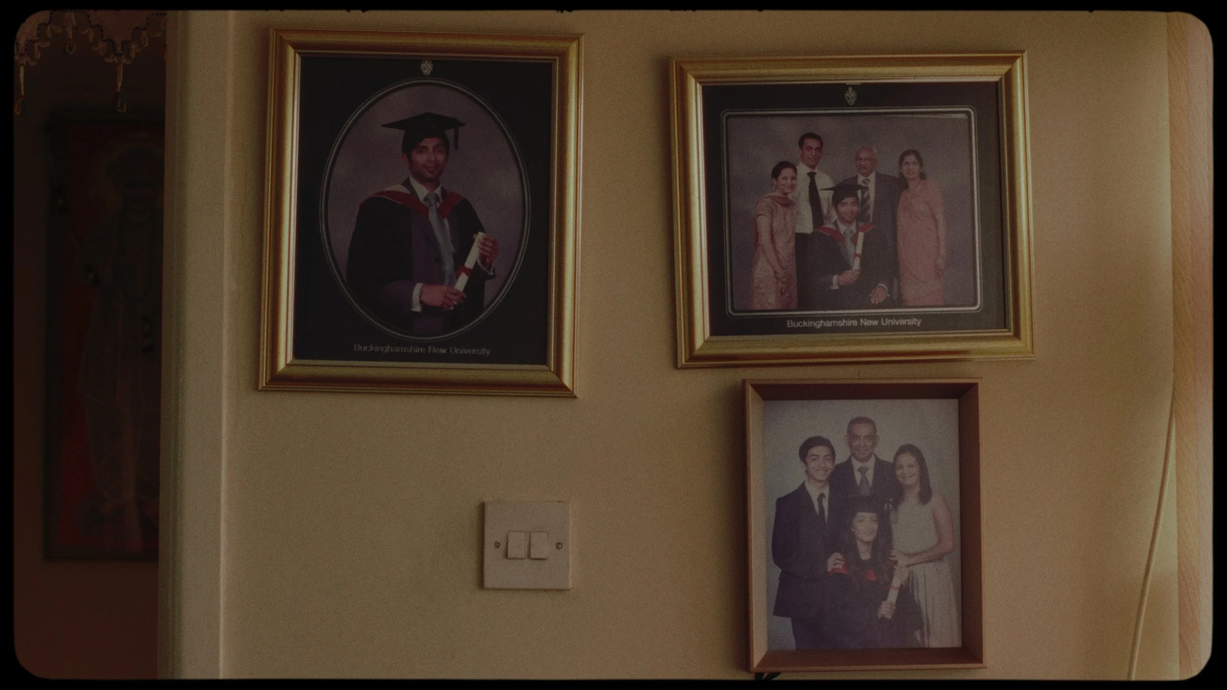 Three framed photographs on a beige wall. The top left photo is of a young man in graduation attire holding a diploma, with the caption 'Buckinghamshire New University.' The top right photo is of a group of five people, also with the same caption. Th