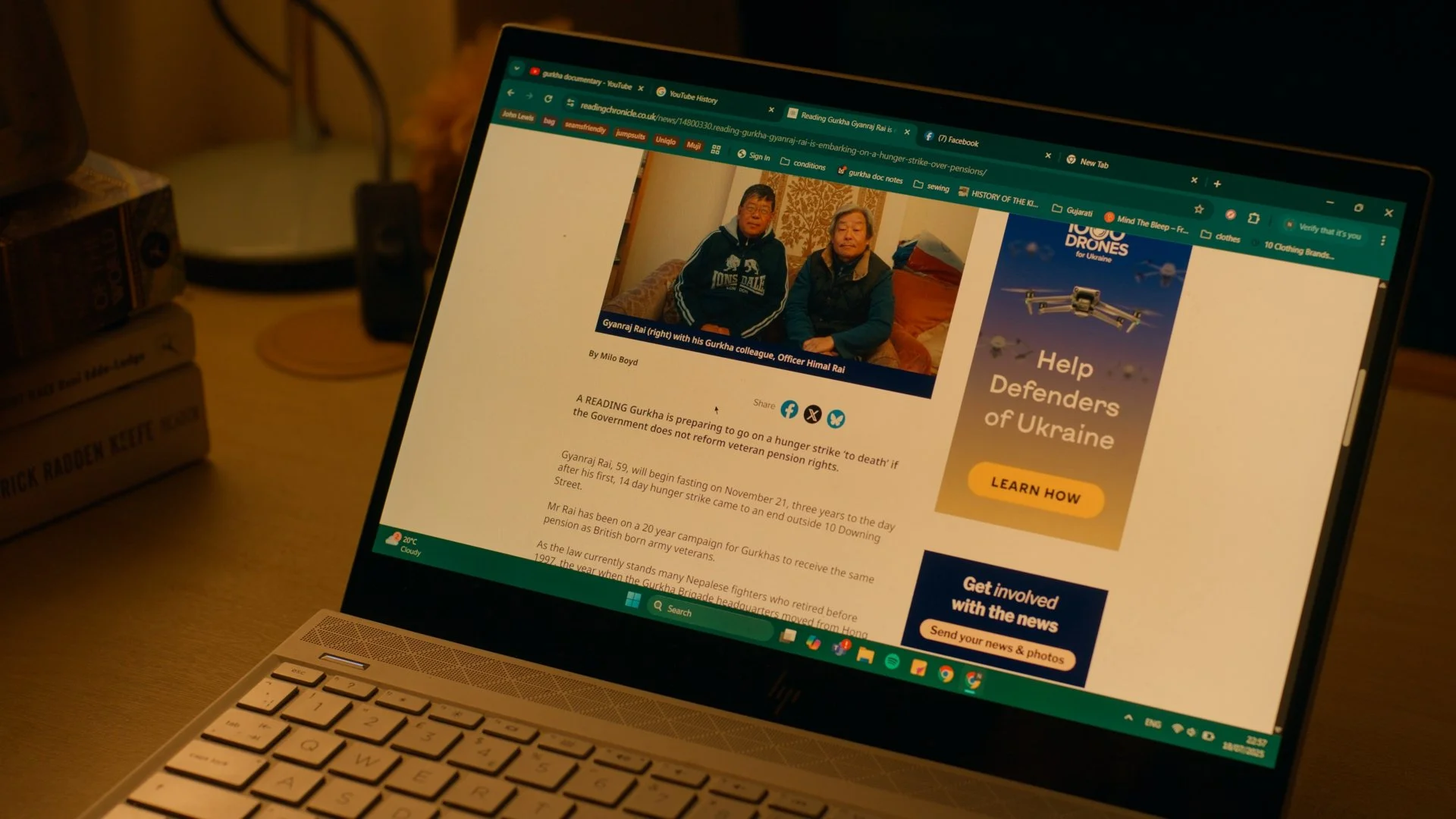 Laptop screen displaying a news article with a photo of two men sitting on a sofa, an advertisement about helping Ukraine, and various tabs at the top. The setting is on a wooden desk with some books, a lamp, and a power strip in the background.