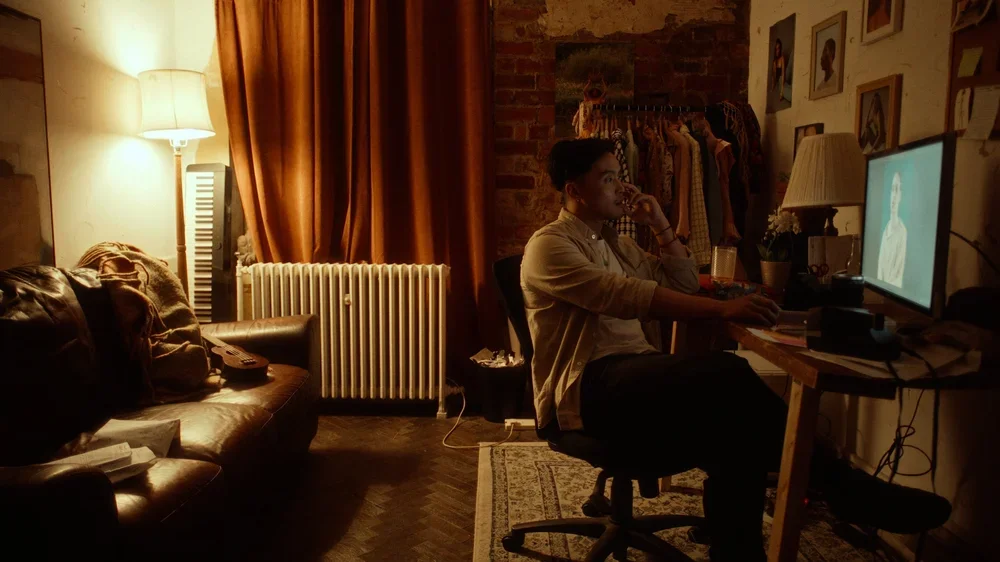 A young man sitting at a desk in a dimly lit room watching a computer screen with a woman on it, surrounded by personal items and framed photographs, with a sofa, a lamp, and curtains in the background.
