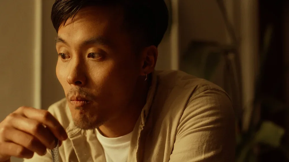 A man with short dark hair, wearing a beige shirt and white t-shirt, sitting at a table in a dimly lit room, eating or drinking something.