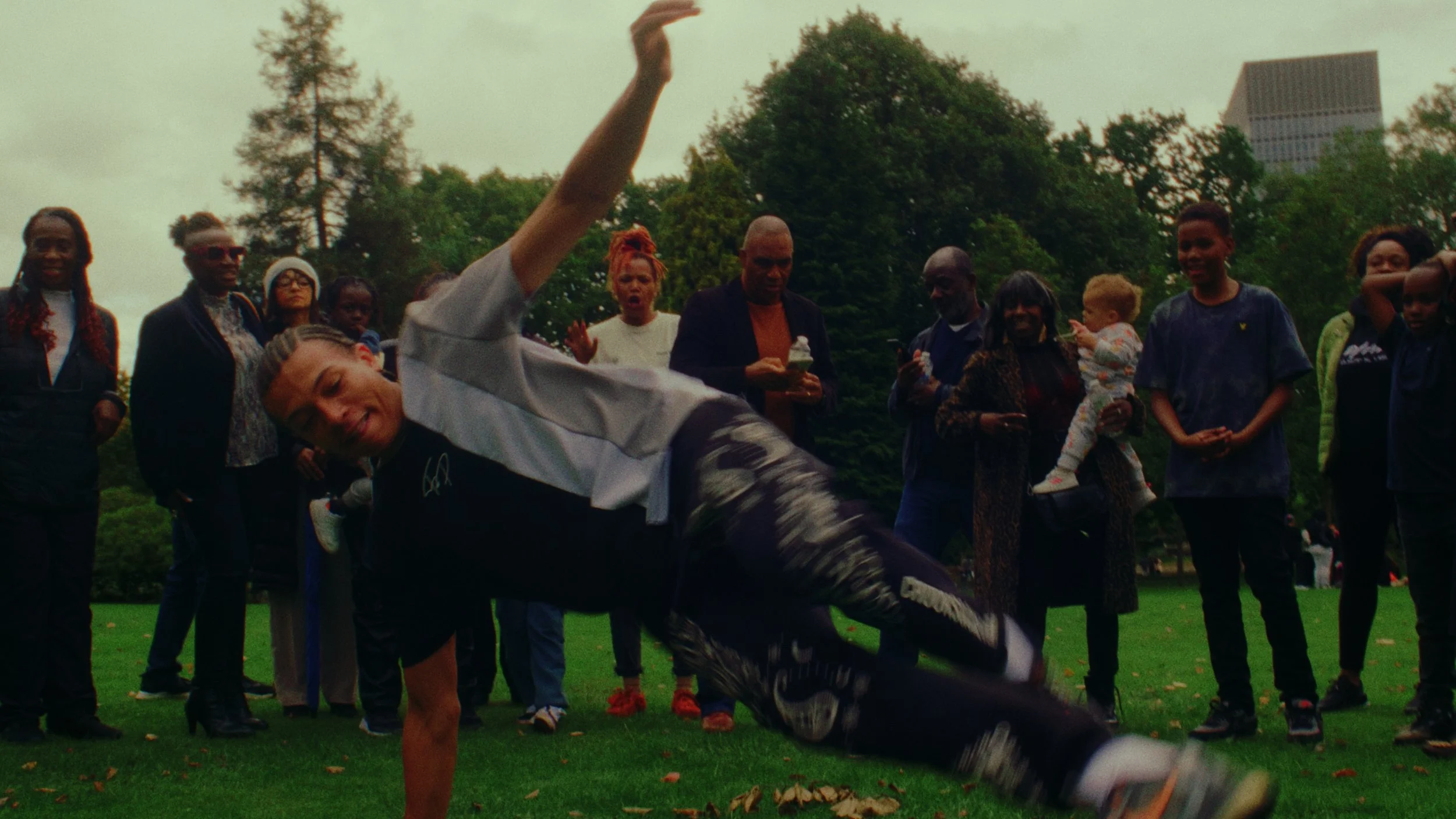 A person in a white and gray jacket performs a breakdance move on the grass while a diverse group of onlookers watches, some with phones, in a park with trees and buildings in the background.