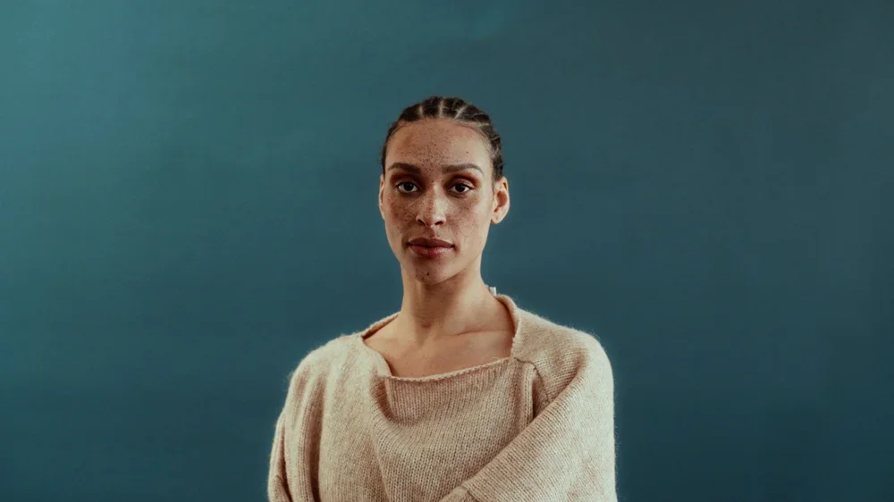 A woman with brown hair in braids wearing a beige sweater stands against a blue background.