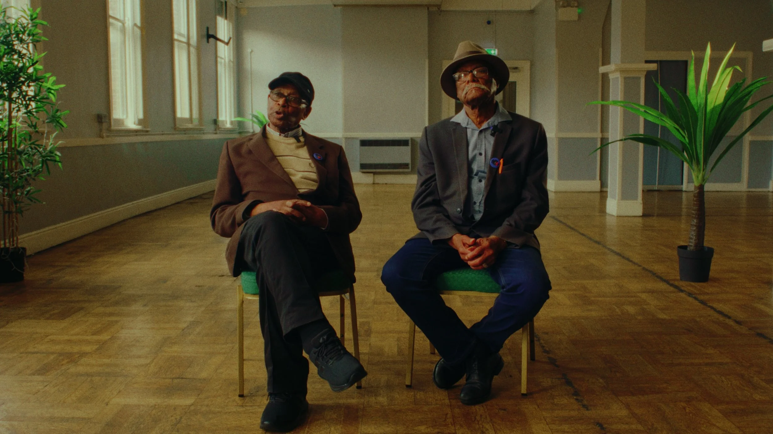 Two elderly men sitting on chairs in a spacious room with wooden floor, large windows, and potted plants.