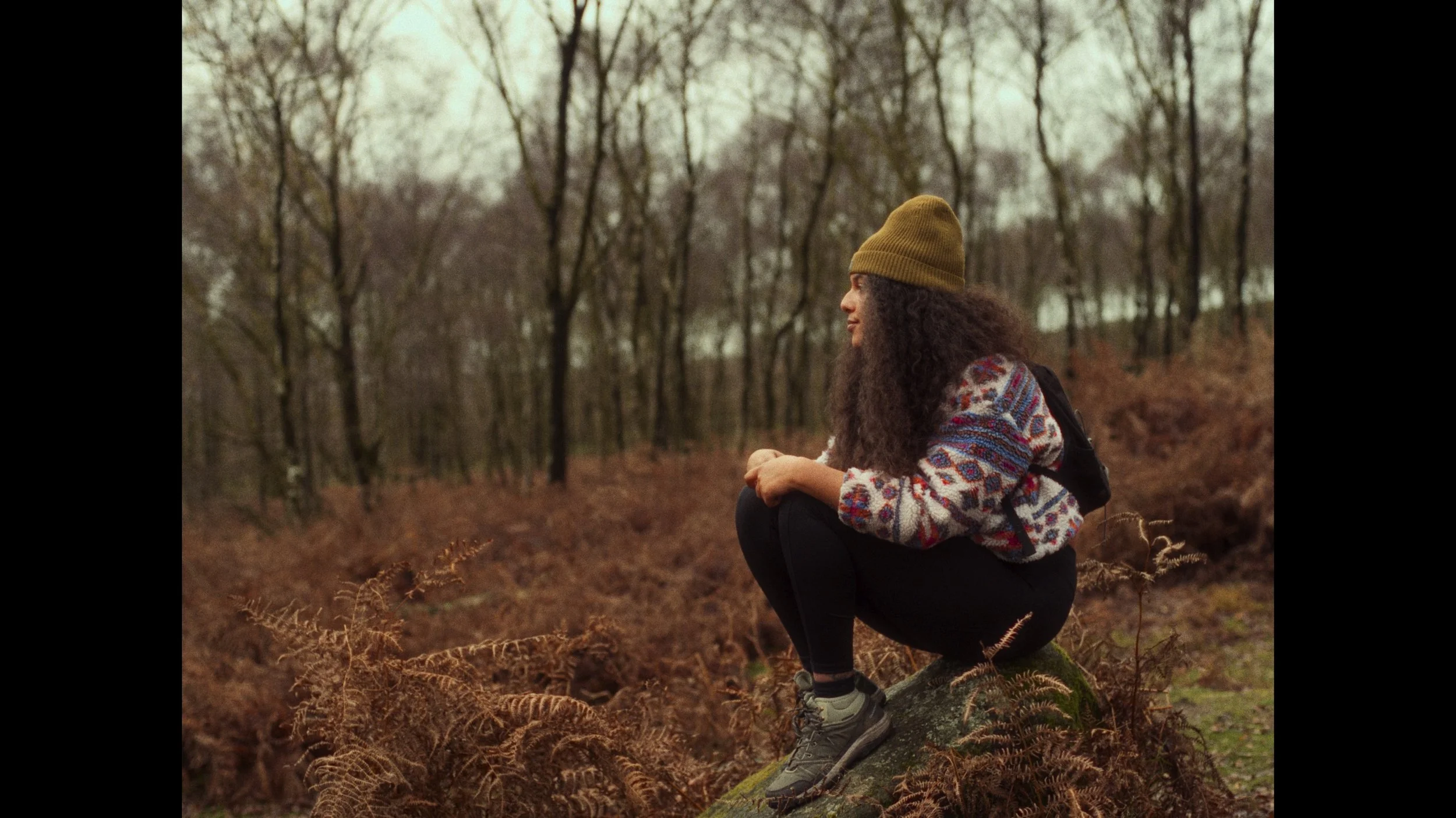 A woman wearing a green beanie, colorful sweater, black pants, and hiking shoes squatting on a log in a forest during autumn, with leafless trees in the background.