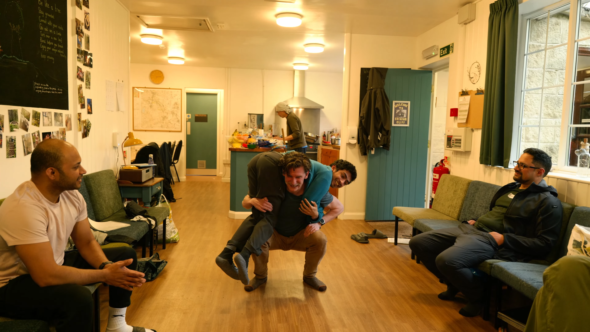 Four men are in a room, one is on another's back in a playful piggyback pose. One man is seated on the left, clapping, another man on the far right is sitting with his hands in his pockets. In the background, a woman is preparing food in the kitchen 