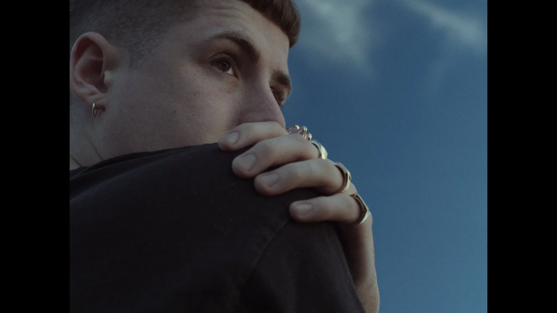 A close-up of a person with short, reddish hair and earrings, resting their head on their hand against a blue sky backdrop.
