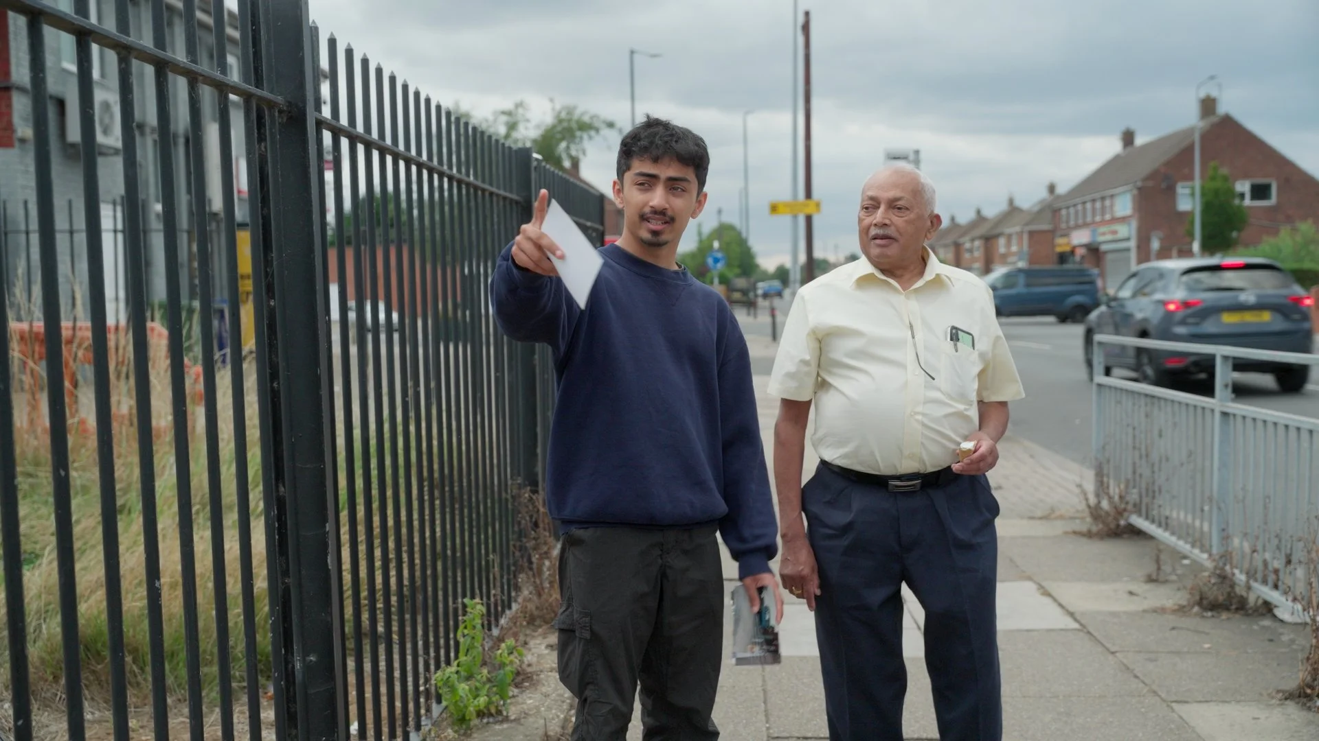 Two men walking on the sidewalk near a black fence, with residential houses and cars in the background. One man points, and the other looks on.